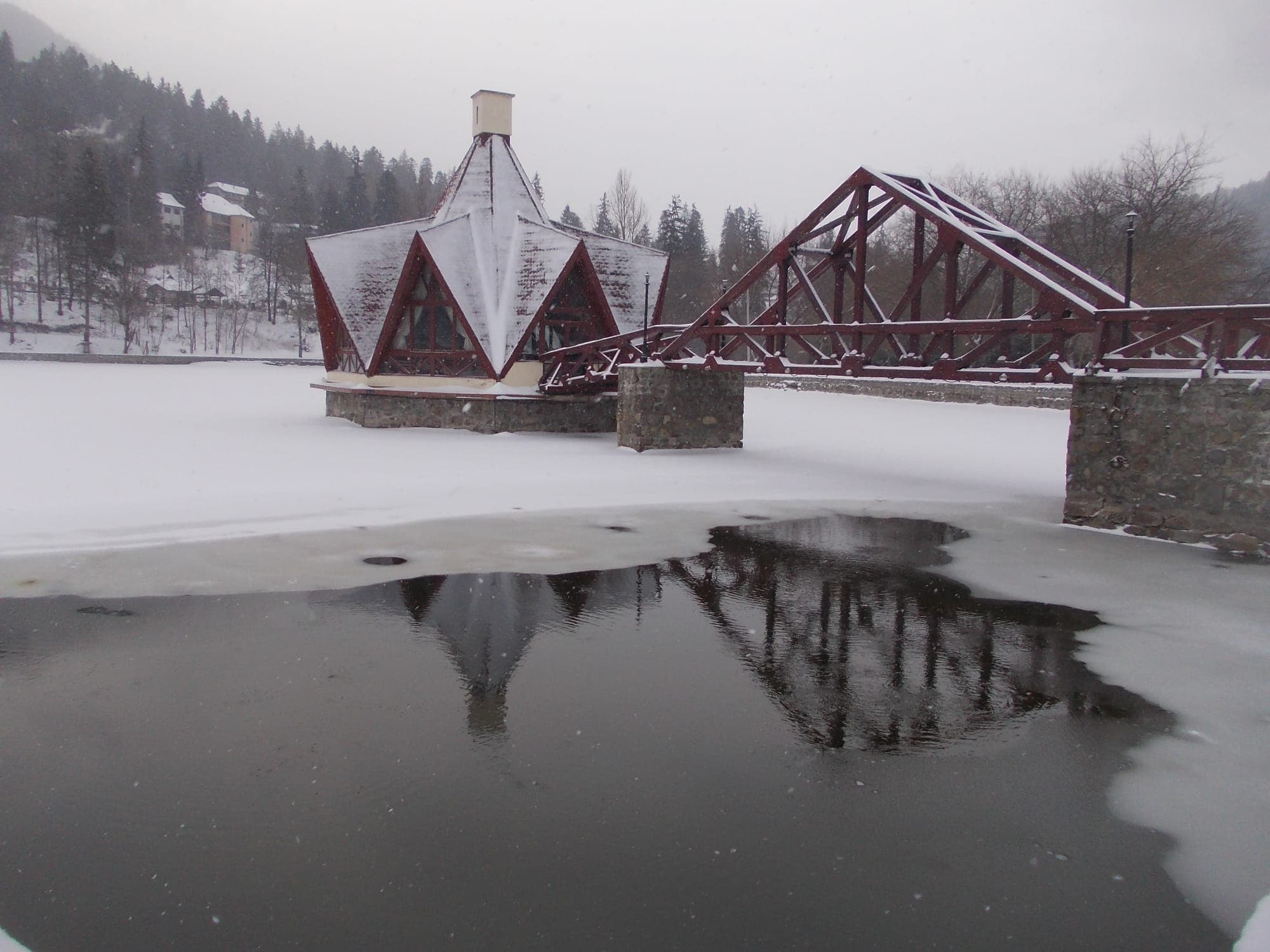 Pavilionul cu acoperiș ascuțit de pe Lacul Ciucaș și podul de lemn roșu care duce la el, iarna, cu lacul înghețat și ninsoare