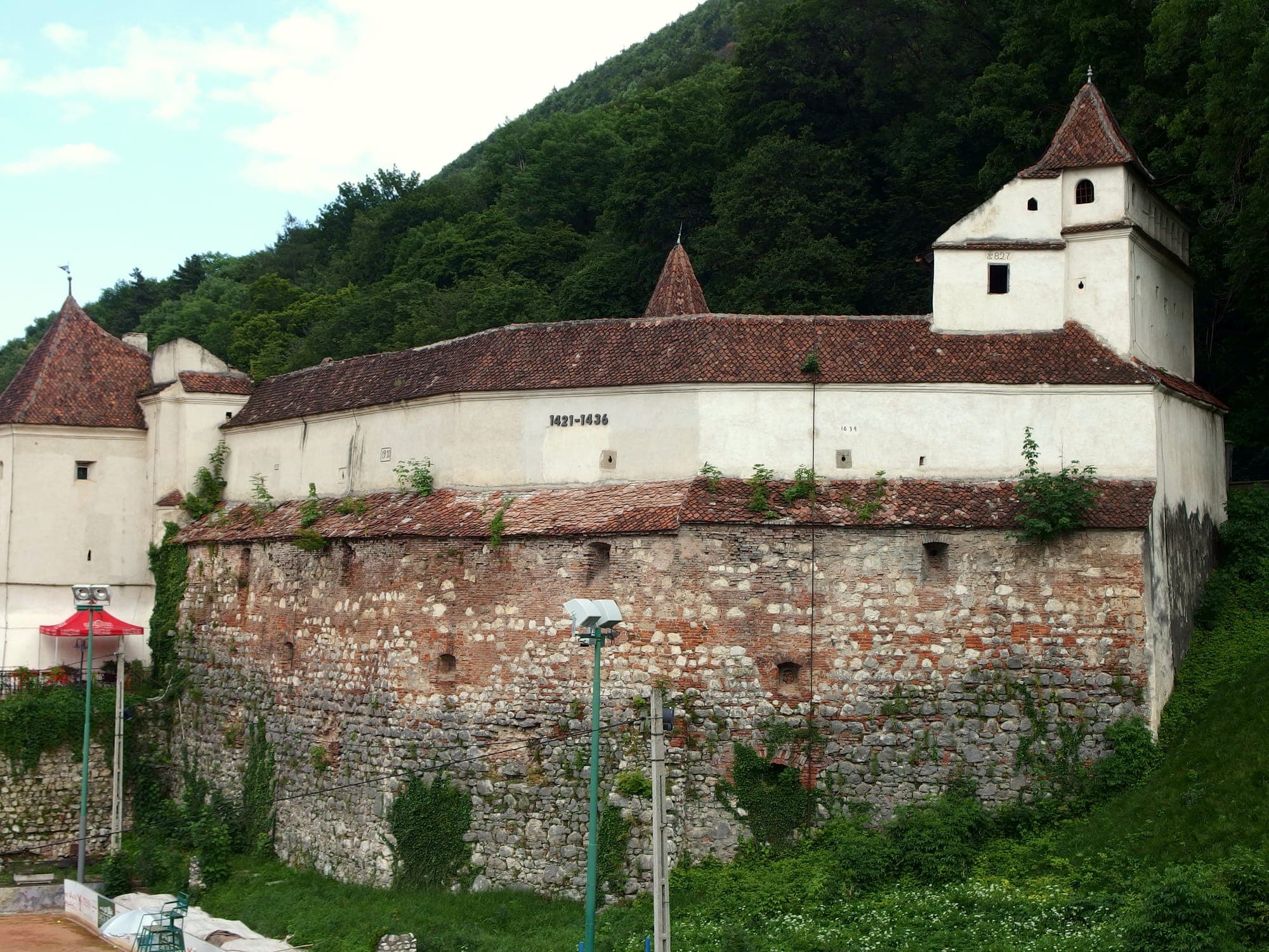 Latura de vest a bastionului, cu zidul de piatră, turnurile cu acoperiș în pantă și pădurea Tâmpei în spate