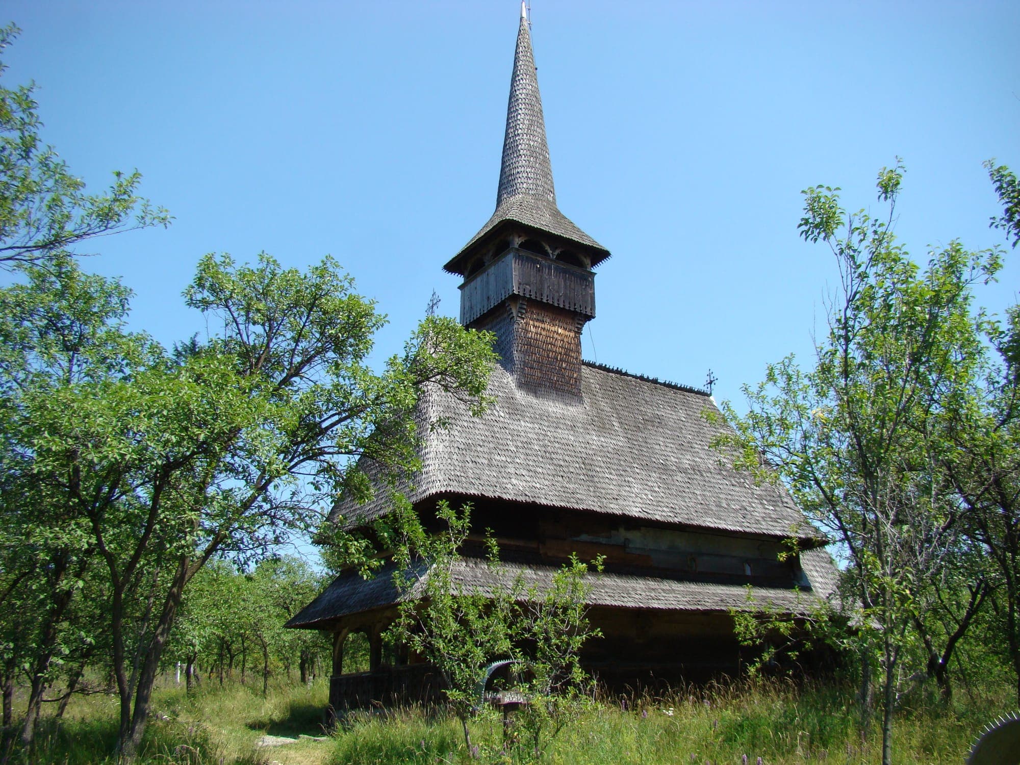Biserica de lemn cu turnul ascuțit acoperit cu șiță, printre pomii livezii de pe deal, vara
