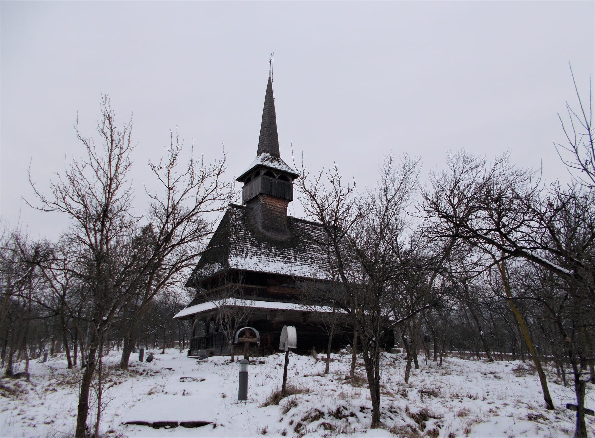 Biserica iarna, cu acoperișul și turnul sub zăpadă, între pomii desfrunziți și crucile cimitirului