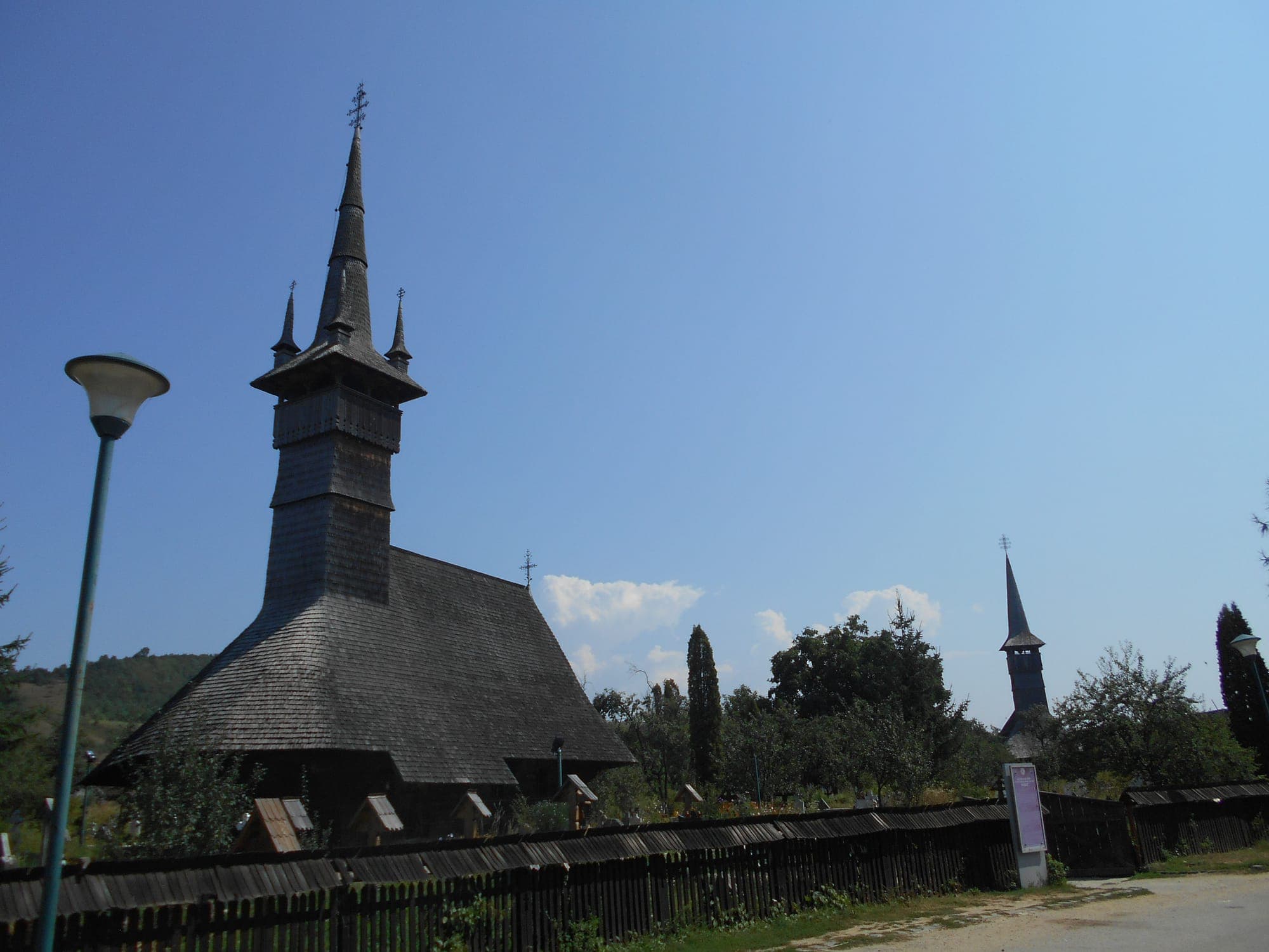 Biserica Sfinții Arhangheli în stânga, după gardul de lemn, iar în depărtare turnul celei de-a doua biserici de lemn din sat