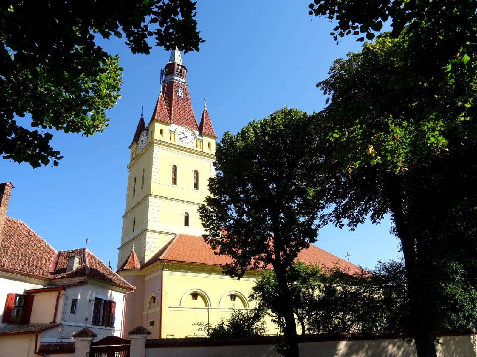Biserica neoclasică galbenă, cu clopotnița și acoperișul de țiglă roșie, văzută printre copaci de dincolo de zid