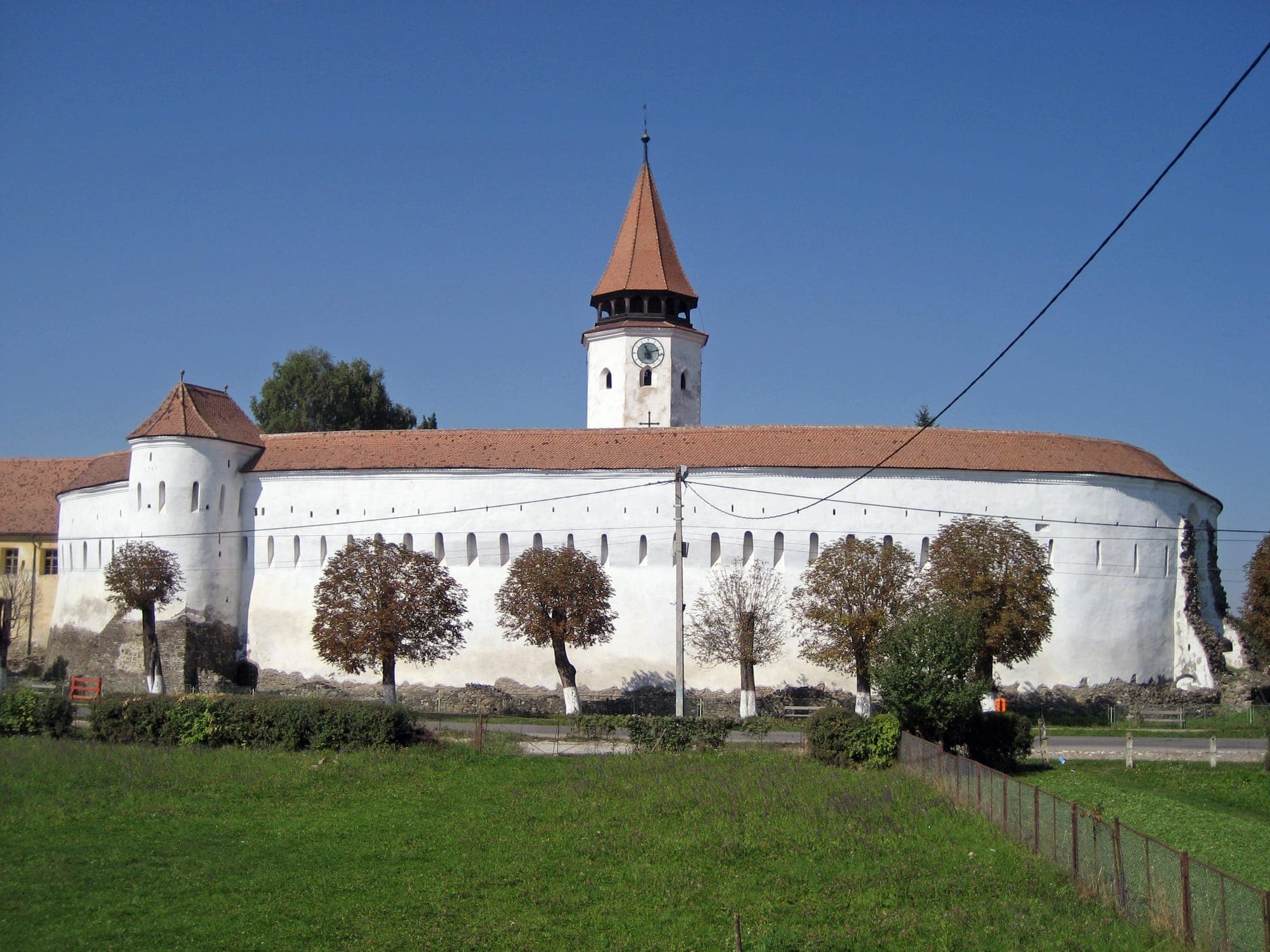 Zidul oval, alb, al cetății cu un turn de colț și turnul cu ceas al bisericii deasupra acoperișului, văzute peste o pajiște