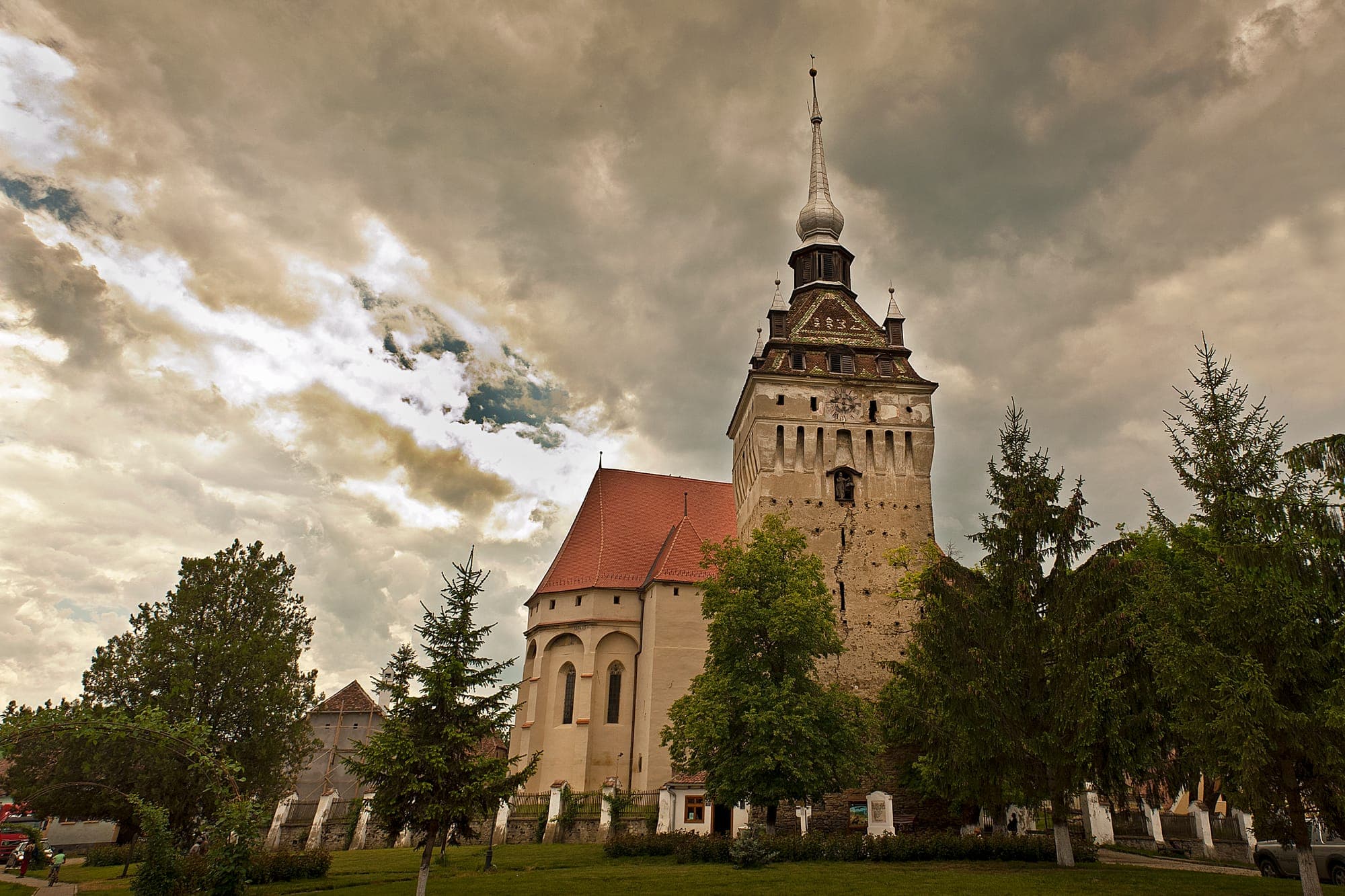 Biserica cu acoperiș roșu și turnul-clopotniță cu coif ascuțit, între brazi, sub un cer înnorat