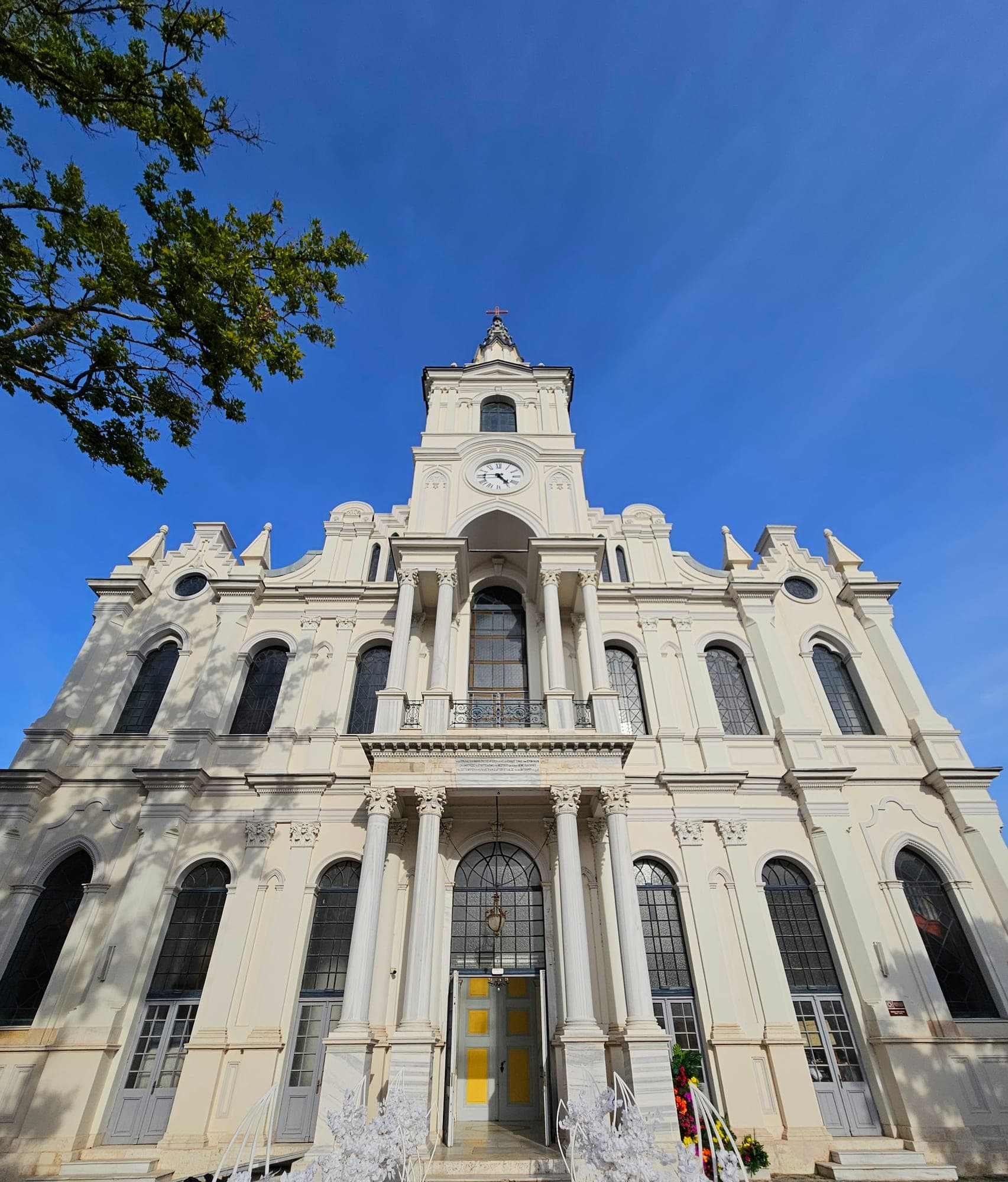 Die Hauptfassade der Kirche von unten gesehen: die Vorhalle mit vier Säulen, der Balkon und der Uhrturm unter blauem Himmel