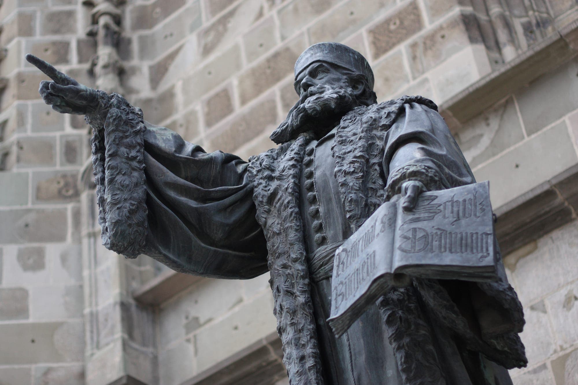 Das Bronzedenkmal von Johannes Honterus mit ausgestrecktem Arm und einem aufgeschlagenen Buch in der Hand, vor der Steinmauer der Kirche