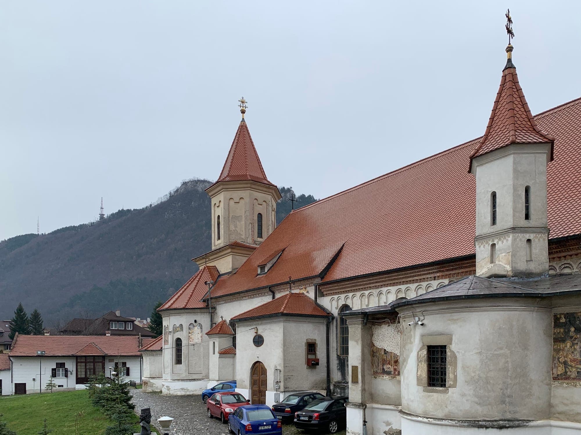 Die Seite der Kirche mit rotem Ziegeldach, Türmchen mit Helmen und bemalter Apsis, im Hintergrund die Zinne (Tâmpa)