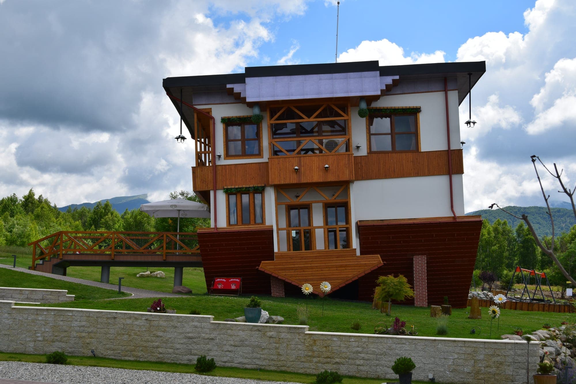 La Casa Capovolta vista di fronte, con il tetto in basso e i piani invertiti, sullo sfondo di bosco e montagne