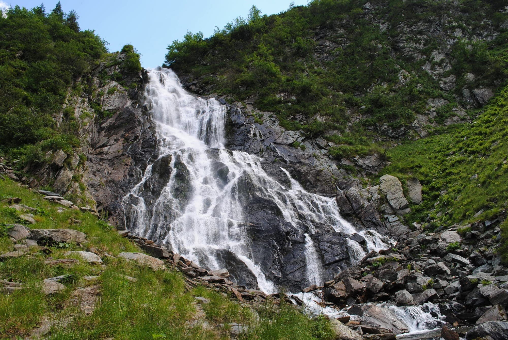 Cascada Bâlea vara, cu apa despletită în șuvițe albe pe peretele de stâncă cenușie, între versanți verzi, văzută de la piciorul ei