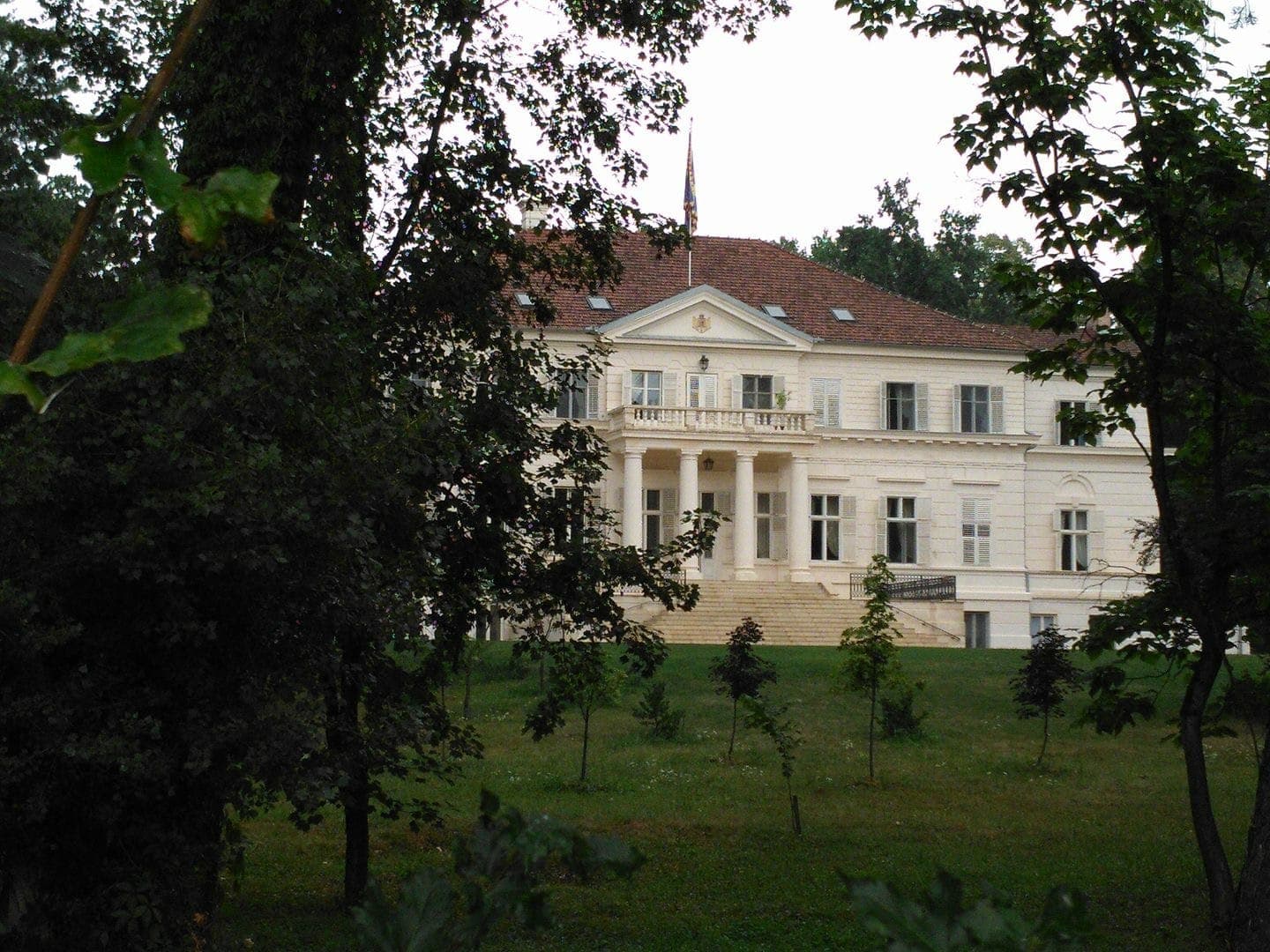 Fațada albă a castelului, cu portic cu coloane, balcon și fronton, văzută printre copaci peste peluza din față
