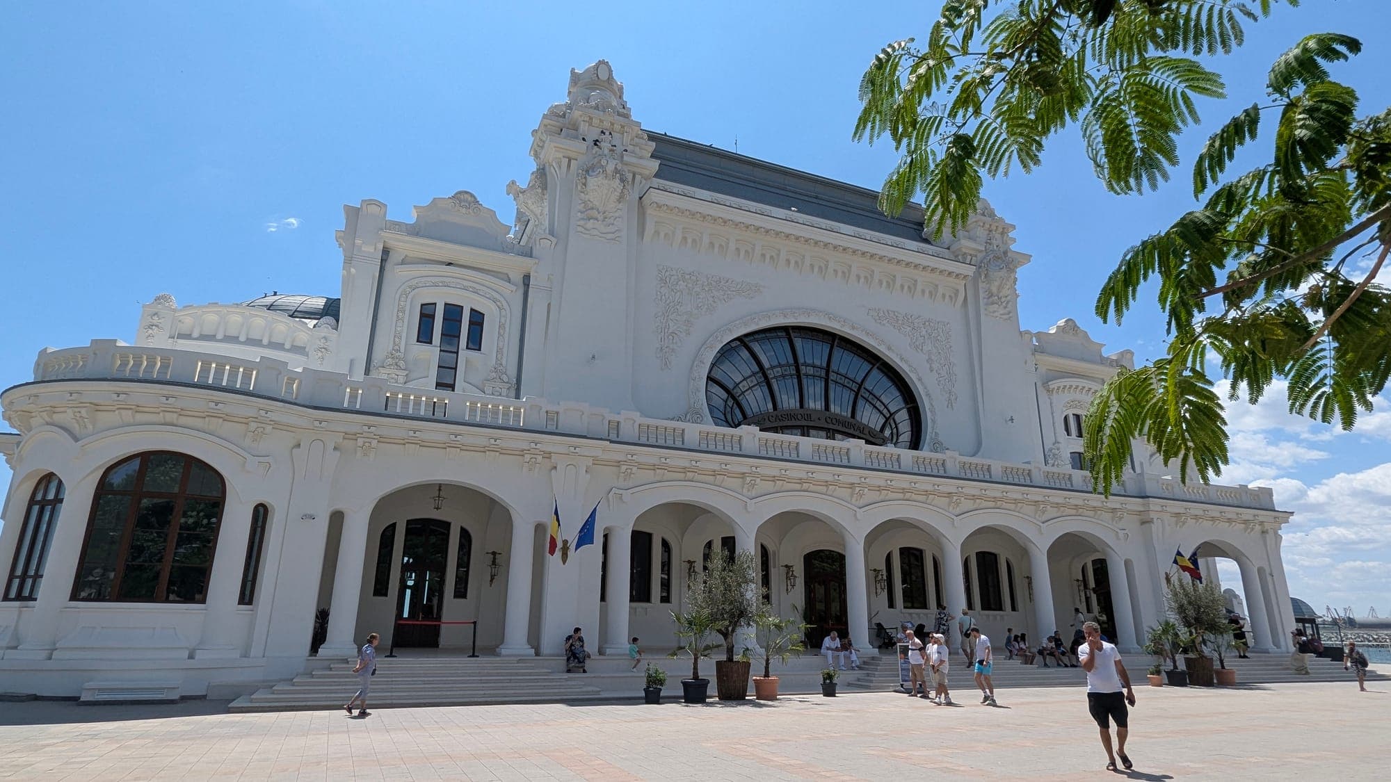 Fațada albă restaurată a Cazinoului dinspre bulevard, cu fereastra semicirculară mare și arcadele de la intrare, sub cer senin