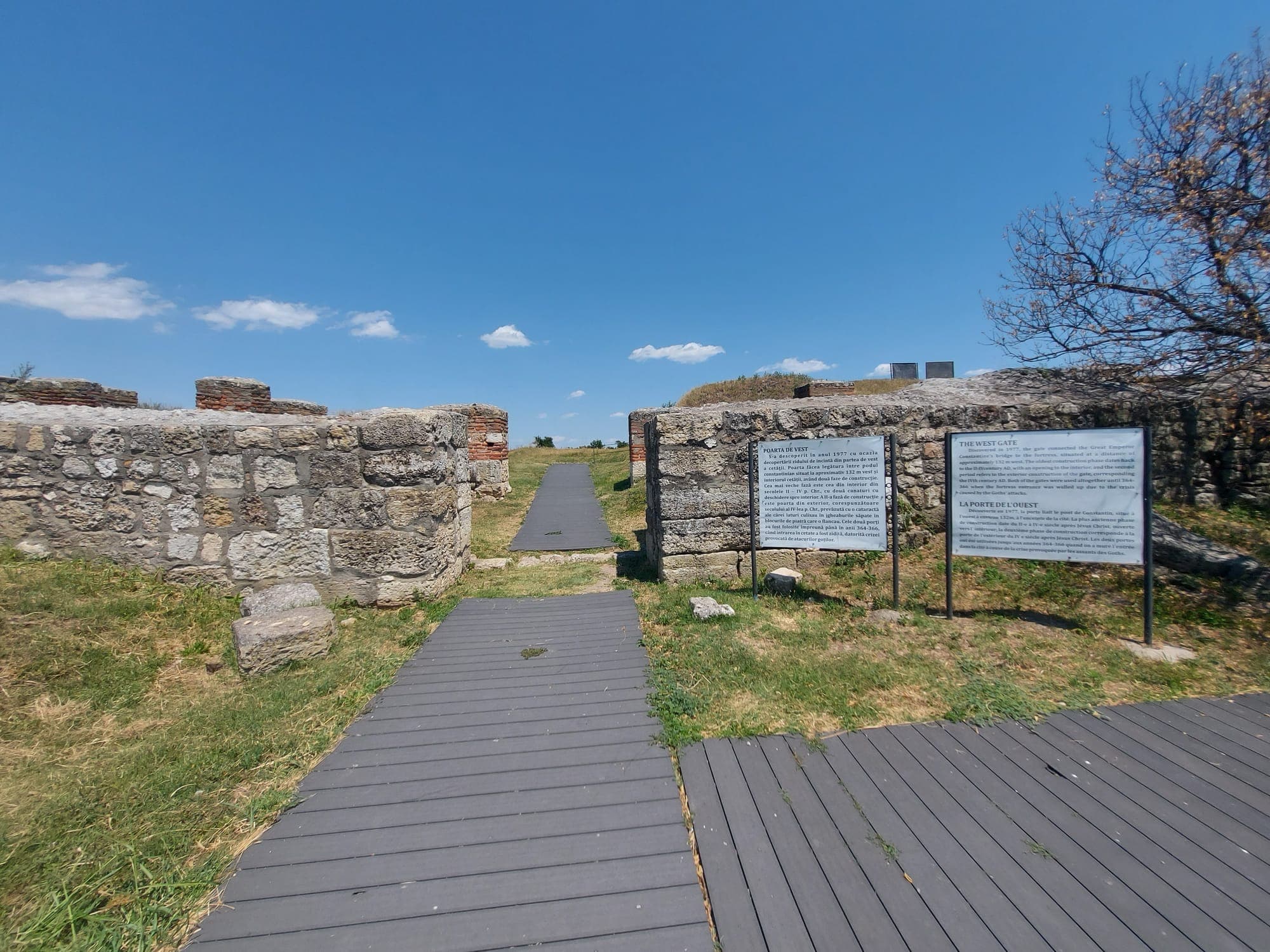 Das Westtor der Festung, mit Steinmauern mit Ziegelbändern, einem Holzsteg und Informationstafeln, unter blauem Himmel