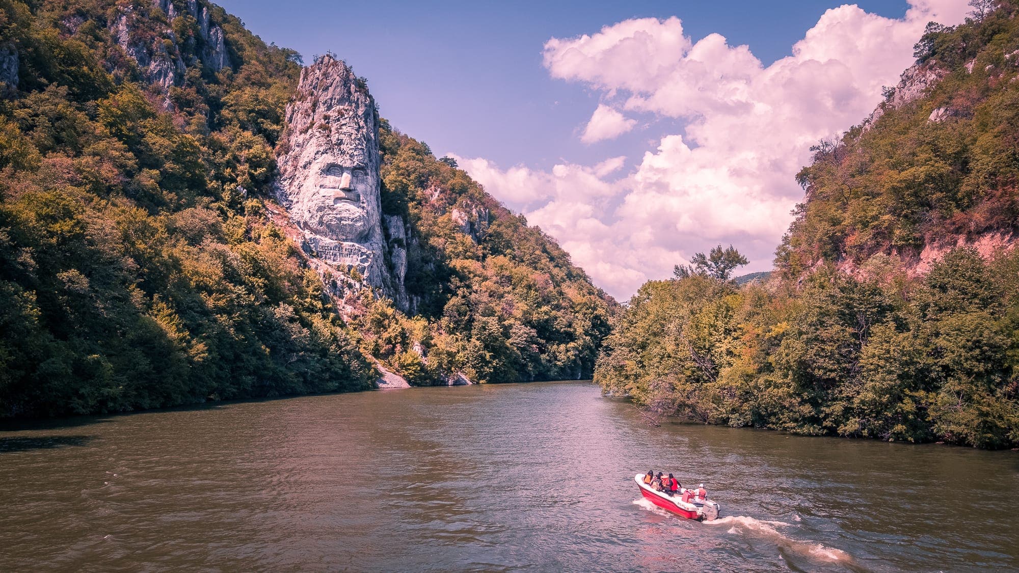 Chipul lui Decebal sculptat în stânca împădurită de deasupra Dunării, cu o barcă cu turiști pe apă, într-o zi senină