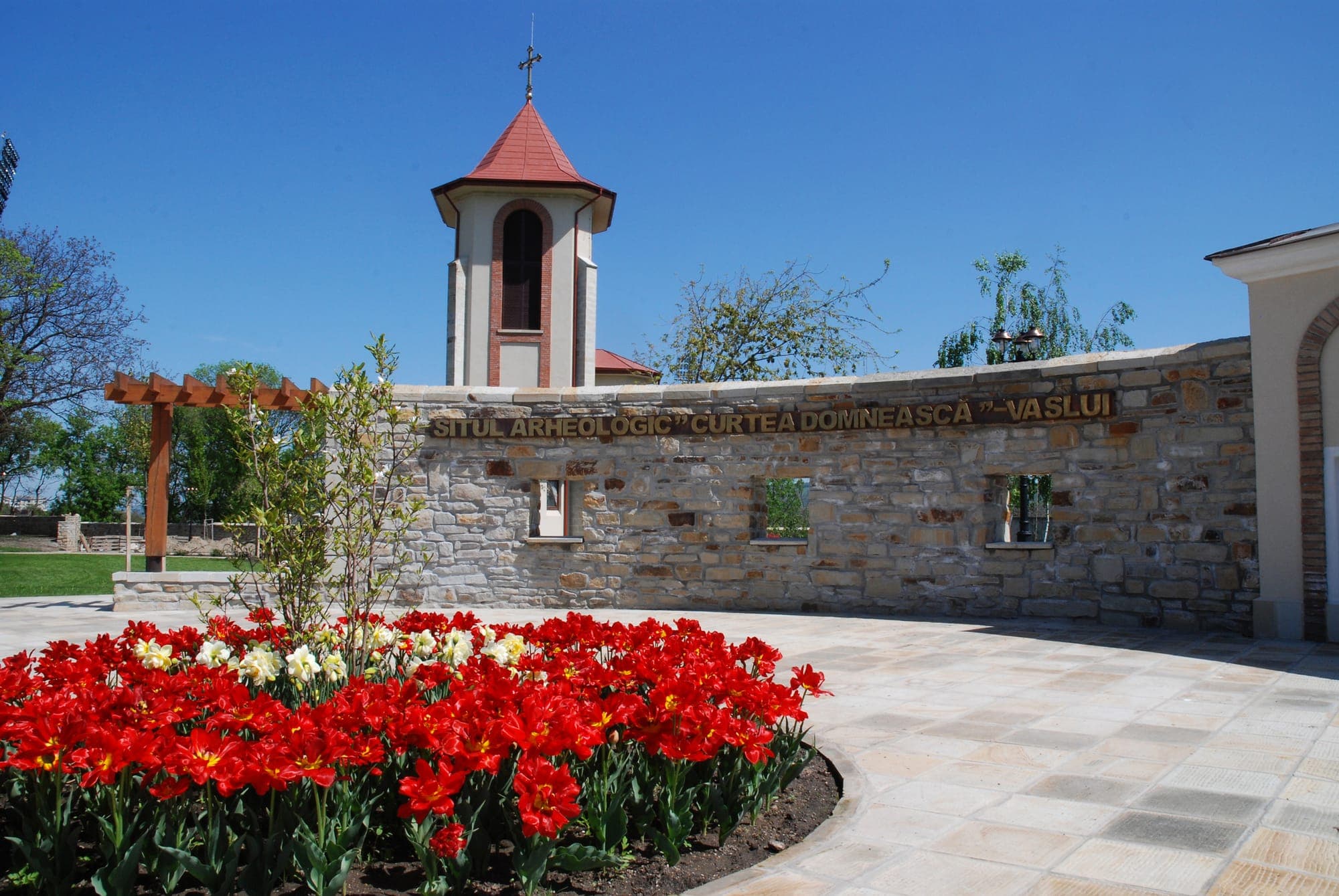 Die Steinmauer mit der Inschrift „Situl arheologic Curtea Domnească Vaslui“, davor Tulpen, dahinter der Glockenturm