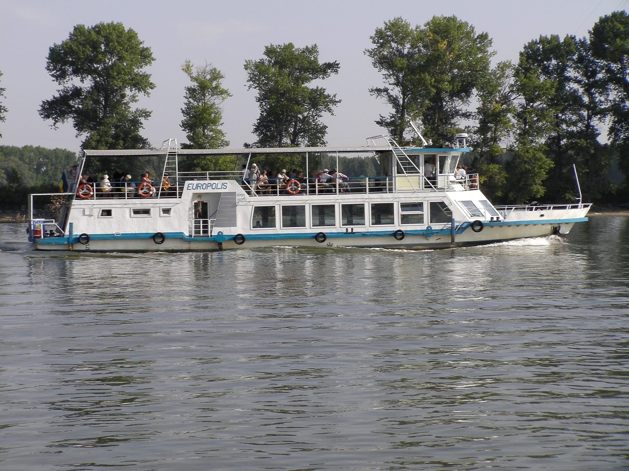 Navă de pasageri cu punte deschisă plină de turiști, pe brațul Dunării, cu plopi pe mal