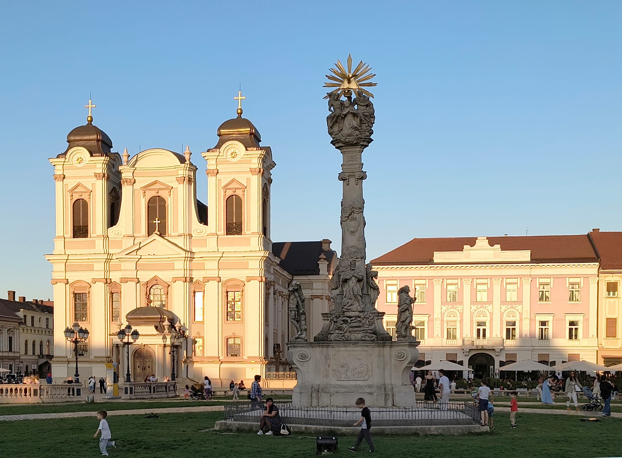 Domul în lumina de seară, cu fațada restaurată în crem și cele două turnuri cu bulbi de cupru, iar în față Monumentul Sfintei Treimi din Piața Unirii