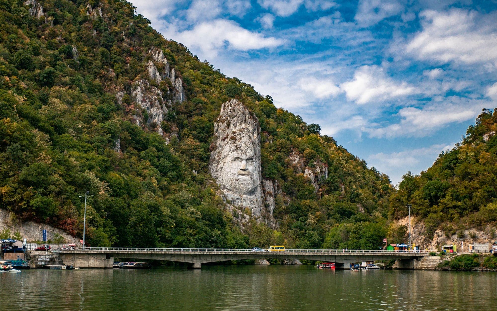 Podul DN57 peste golful Mraconia, cu mașini pe el și chipul lui Decebal sculptat în stânca de deasupra, văzut de pe Dunăre