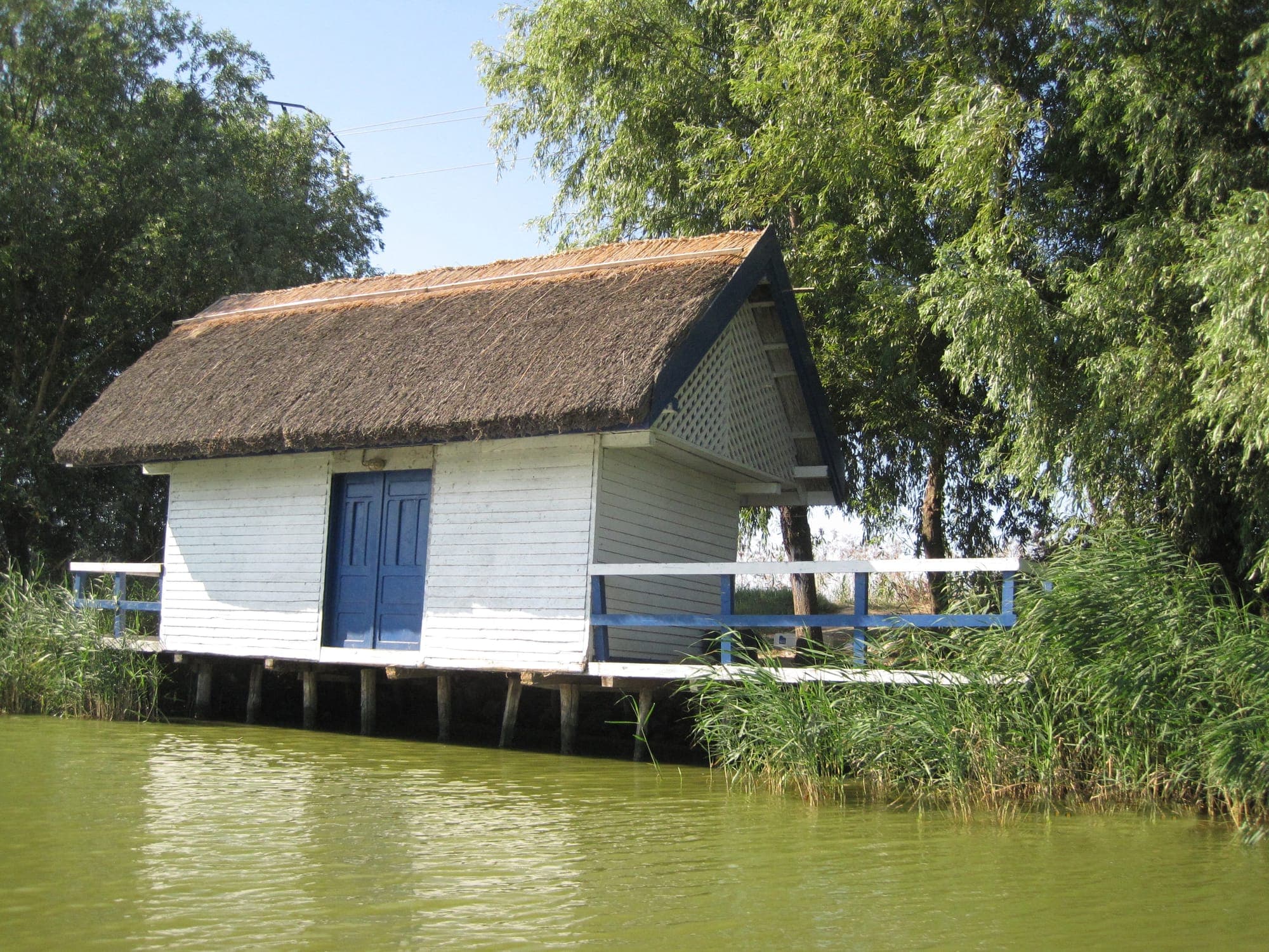 Căsuță albă cu acoperiș de stuf, ridicată pe piloni deasupra apei, între trestii, pe malul dinspre lac