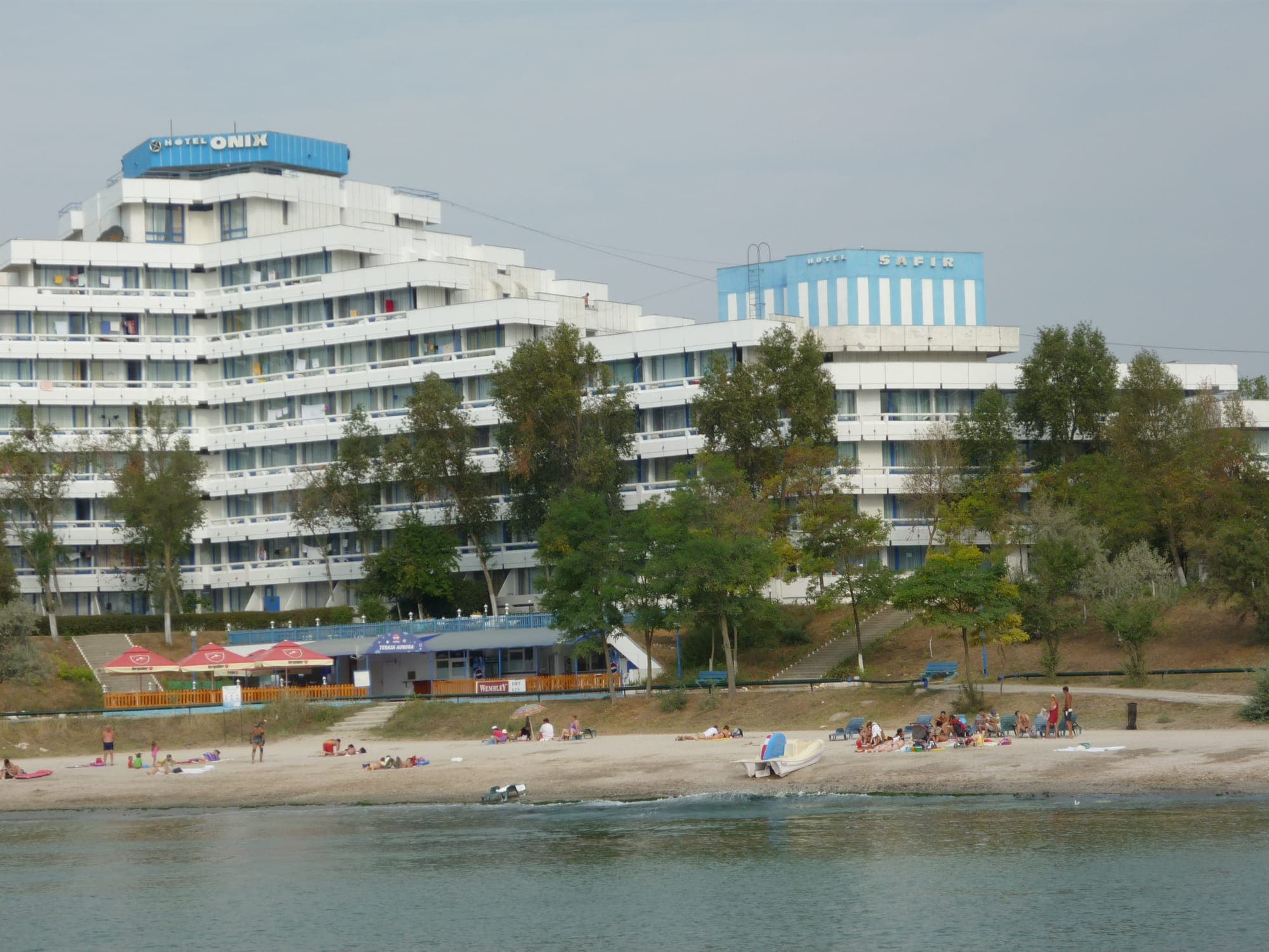 Hotelurile Onix și Safir, albe, cu etajele retrase în trepte, deasupra plajei cu nisip și a unui rând de copaci, văzute dinspre mare