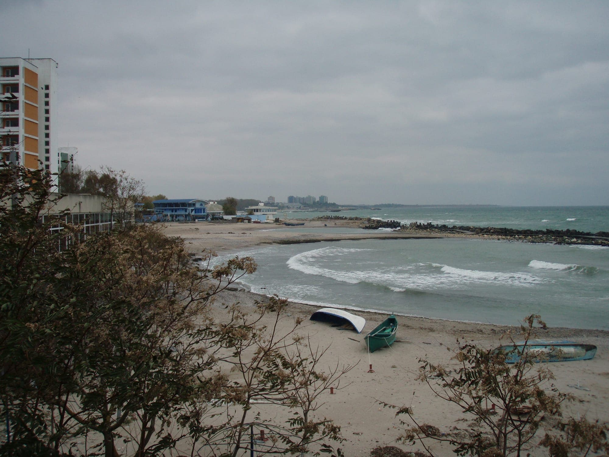 Eine Sandbucht mit an Land gezogenen Booten, von Steinbuhnen abgeschlossen, links ein hohes Hotel und in der Ferne die Türme von Neptun, bei bedecktem Himmel