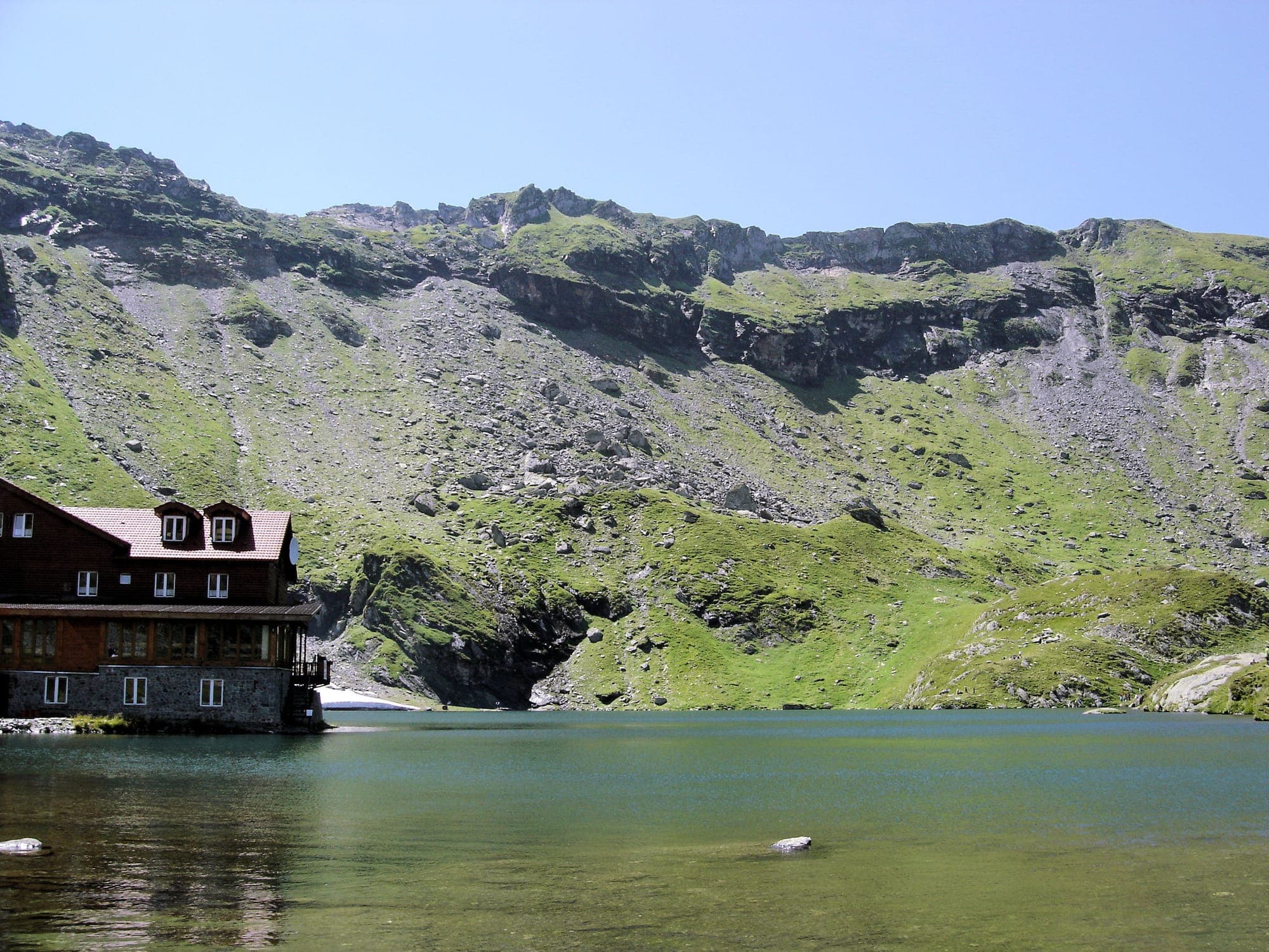 Cabana de lemn și piatră de pe malul lacului, cu apa limpede și versantul înverzit al căldării