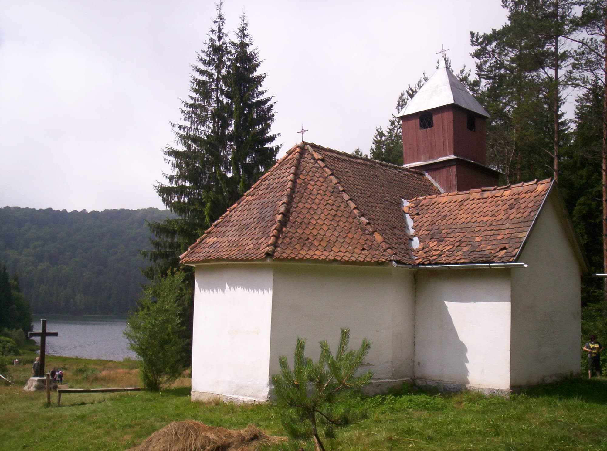 Capela albă a Sfintei Ana, cu acoperiș de țiglă și clopotniță de lemn, între brazi, cu lacul în stânga