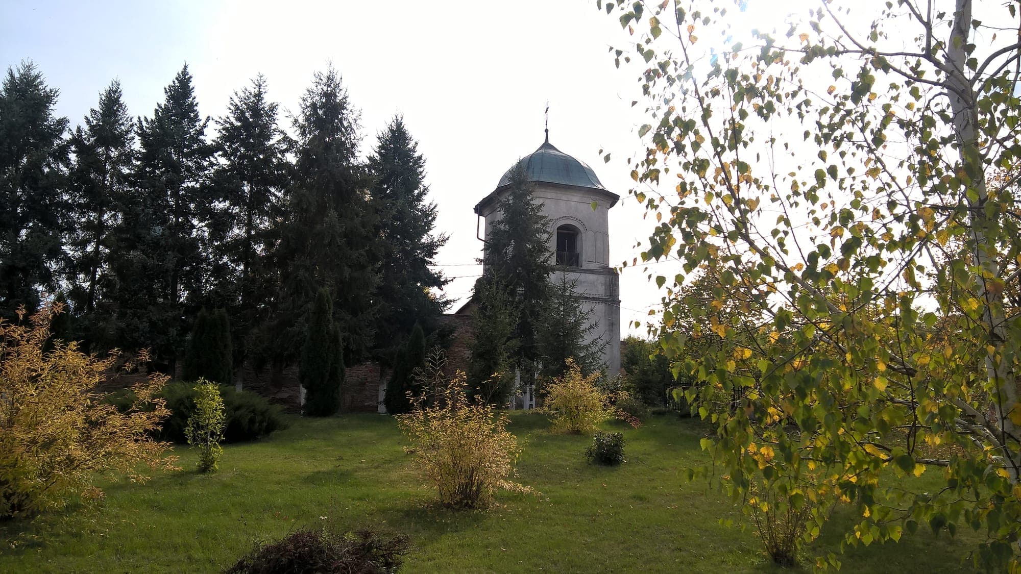 Der Glockenturm mit Torbogen, hinter einer Wiese mit Sträuchern und einer Birke im Herbst