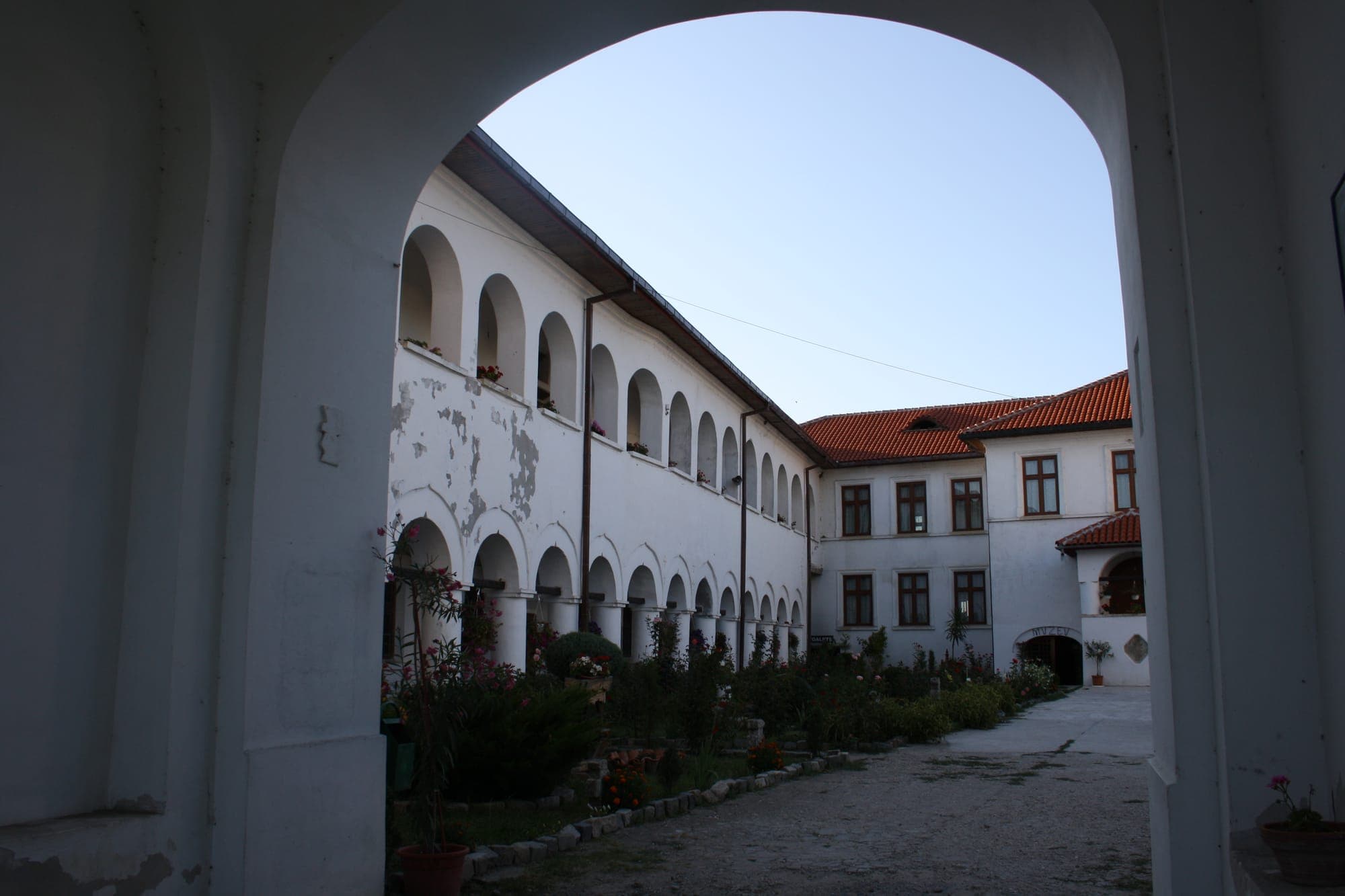 Chiliile cu arcade ale mănăstirii, văzute printr-o boltă a porții, cu flori în curte