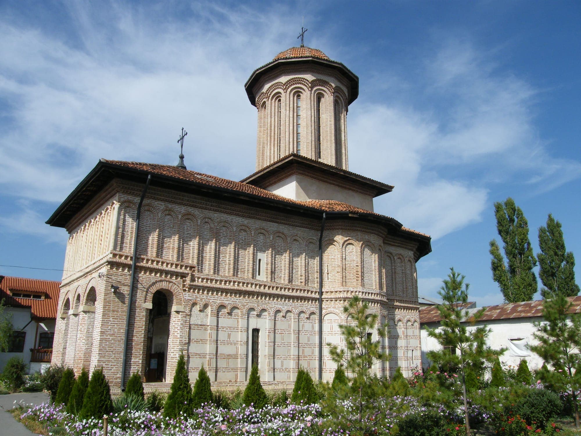 Die Klosterkirche mit ihren Fassaden aus sichtbarem Backstein und dem zentralen Turm, von der Ecke aus gesehen, zwischen Blumen und Thujen unter leicht bewölktem Himmel