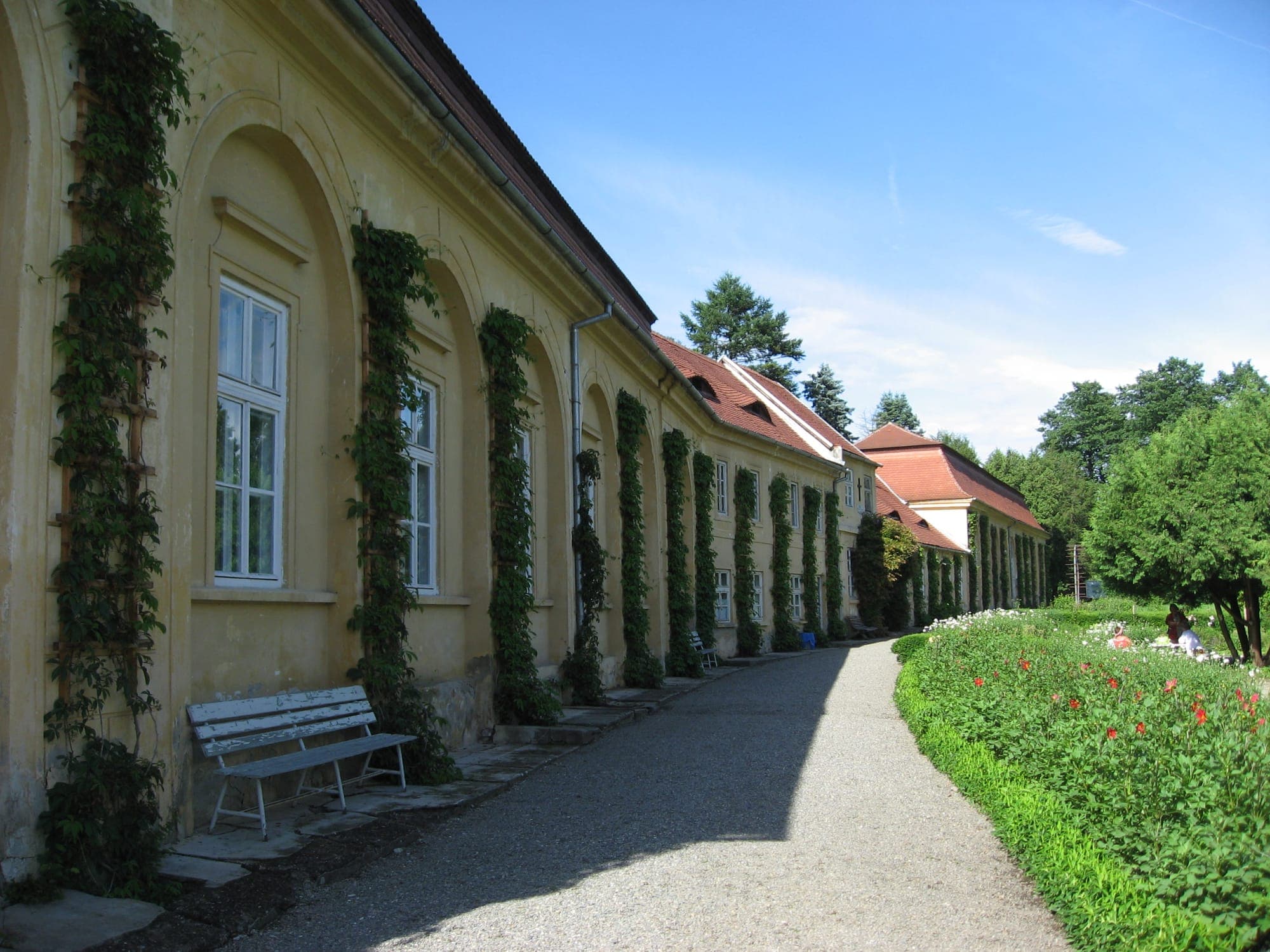 Die Fassade der Orangerie mit hohen Bögen und Kletterpflanzen, neben einem Kiesweg und einem Blumenbeet