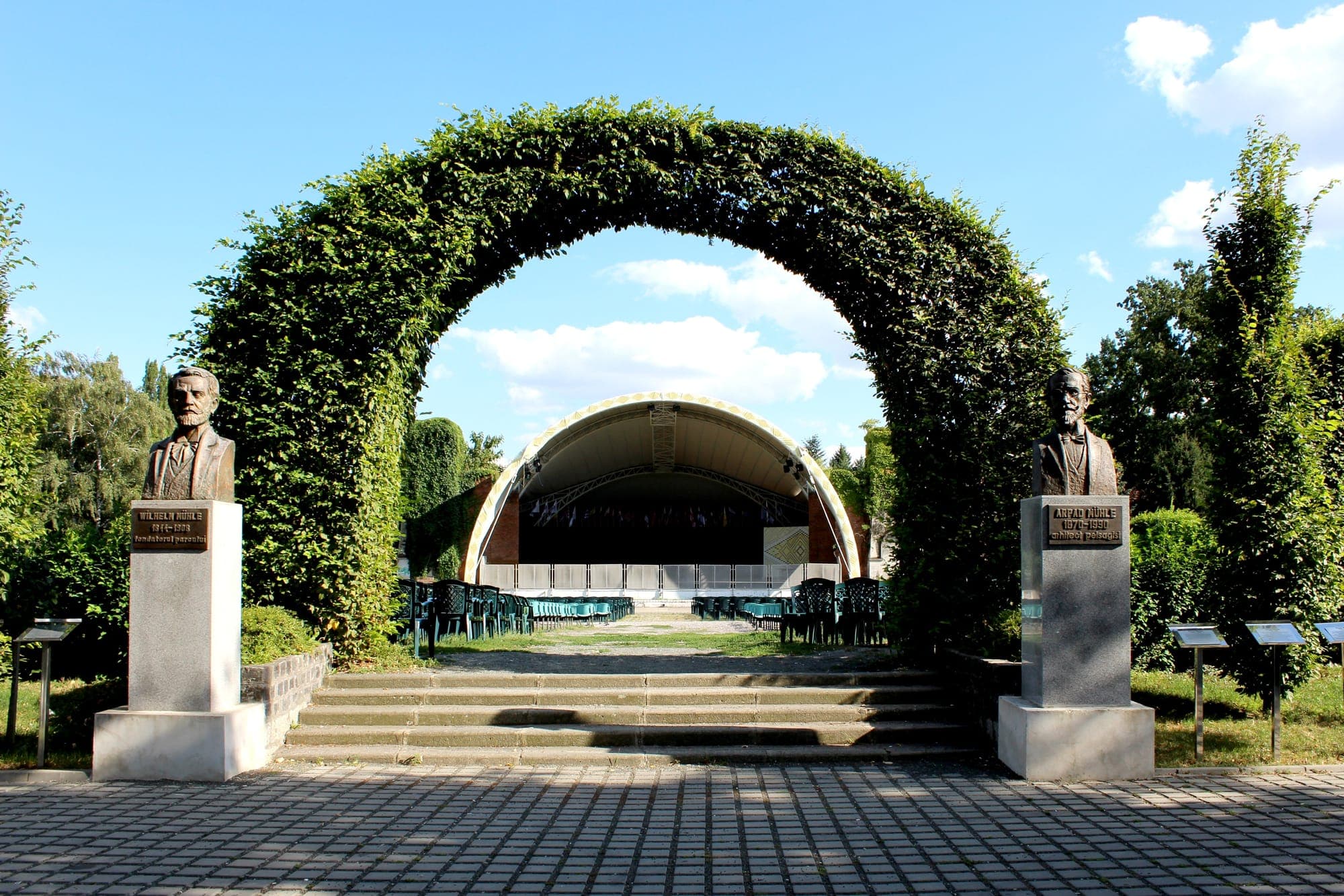 Der Parkeingang, mit den Büsten von Wilhelm Mühle und Árpád Mühle zu beiden Seiten eines Bogens aus Grün, hinter dem die Bühne des Sommertheaters zu sehen ist