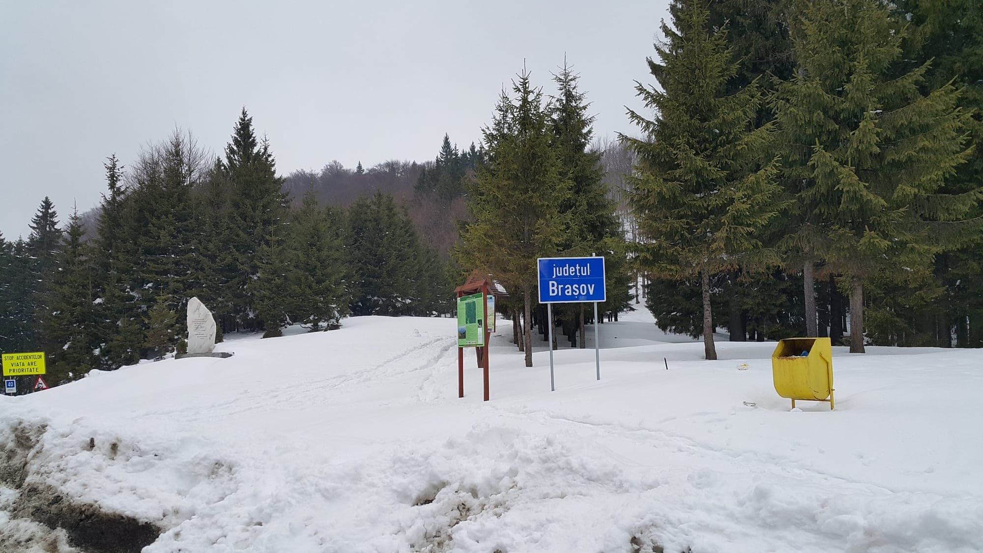 Șaua pasului iarna, acoperită de zăpadă: monumentul de marmură, panoul cu trasee și indicatorul „județul Brașov”, între molizi