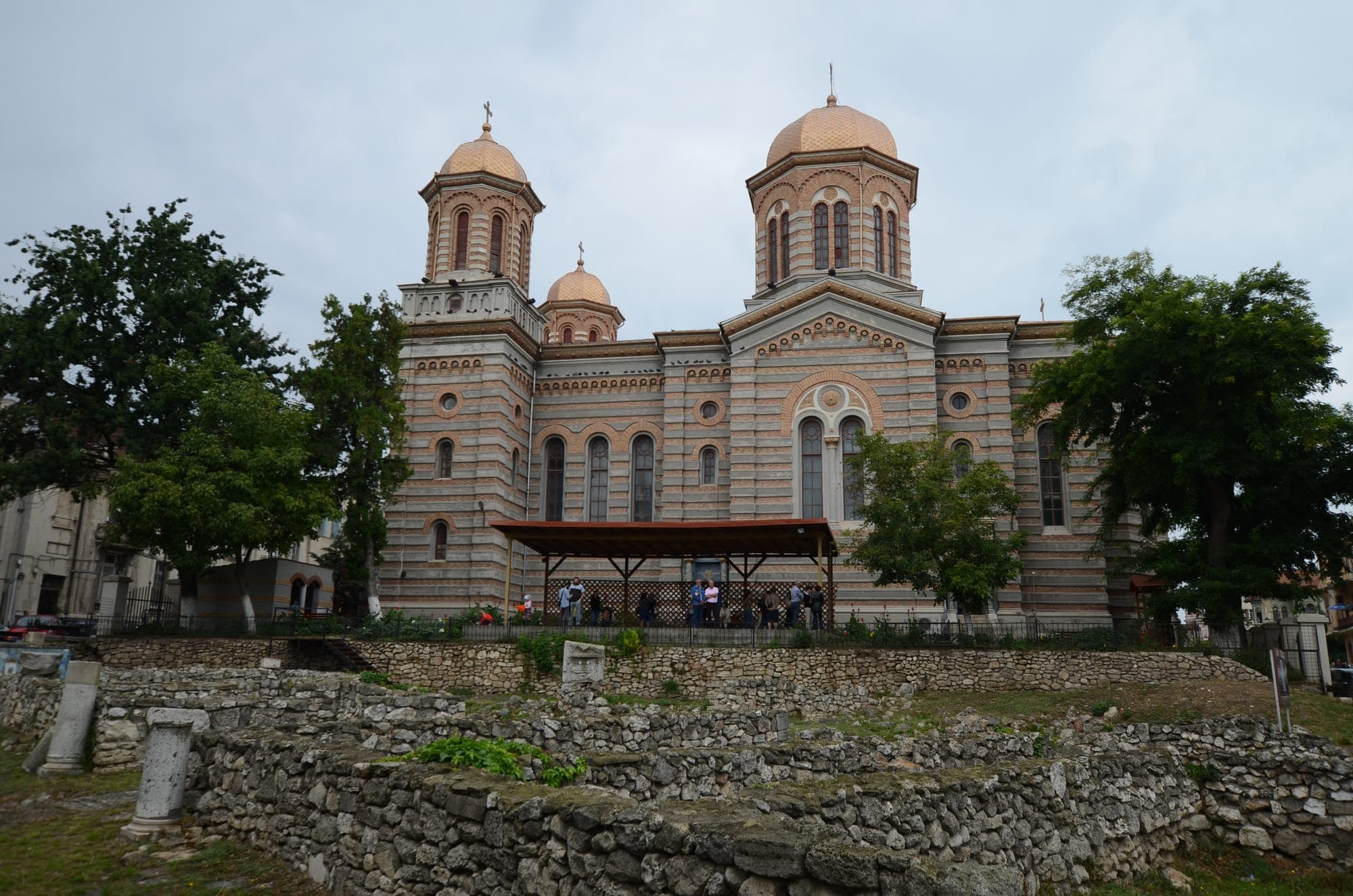 Catedrala Sfinții Petru și Pavel, din cărămidă în dungi, cu turle aurii, văzută peste zidurile antice ale Tomisului din parc