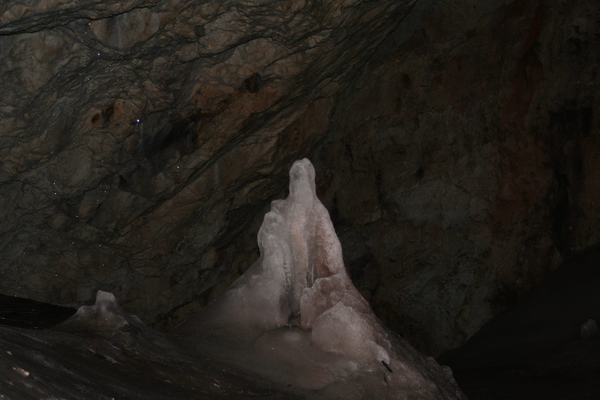 Stalagmită de gheață albă, înaltă cât un om, ridicată din podeaua înghețată a peșterii în fața unui perete întunecat