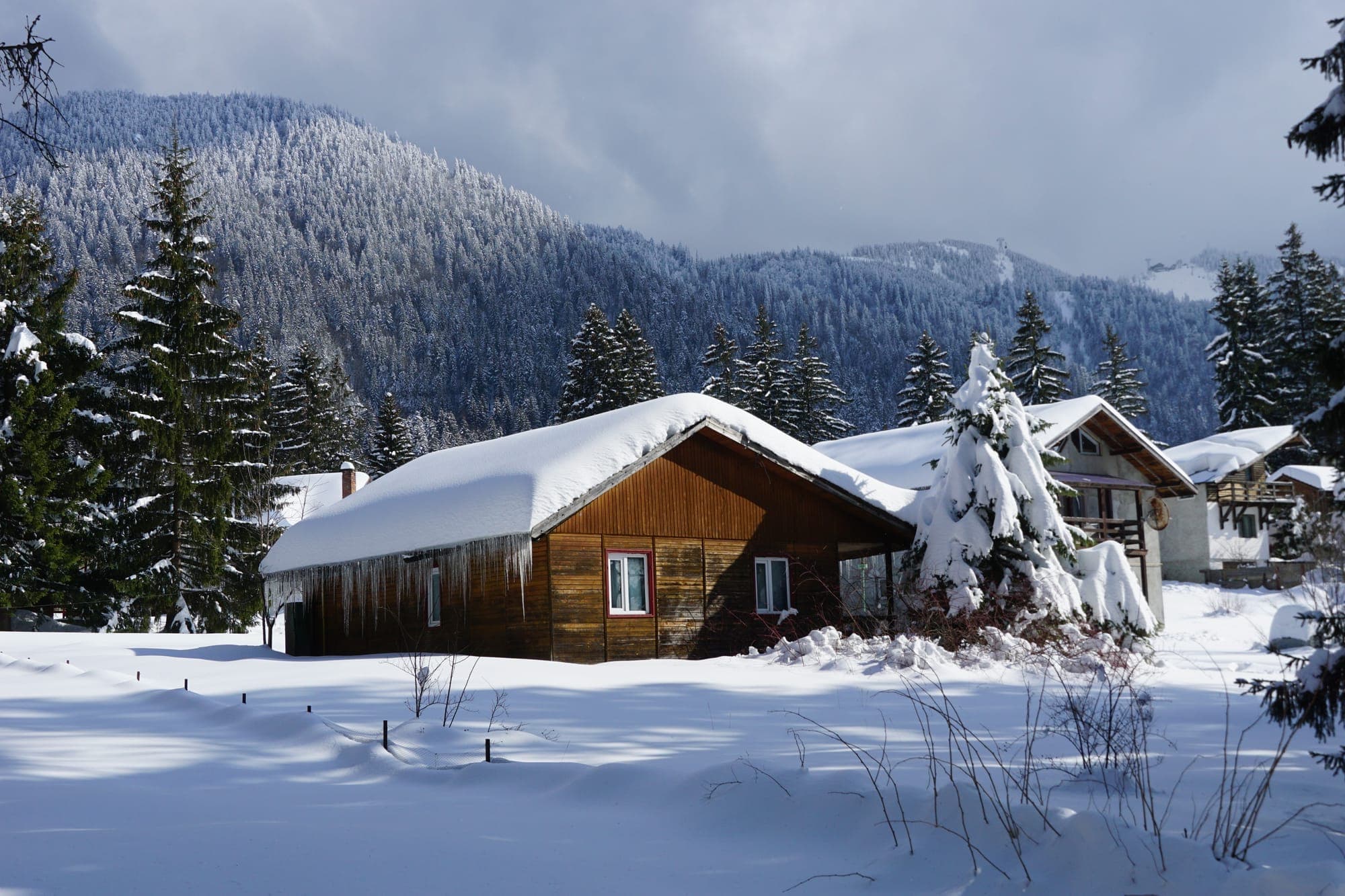 Una baita di legno con il tetto carico di neve e ghiaccioli, sotto i versanti boscosi e bianchi del Postăvaru