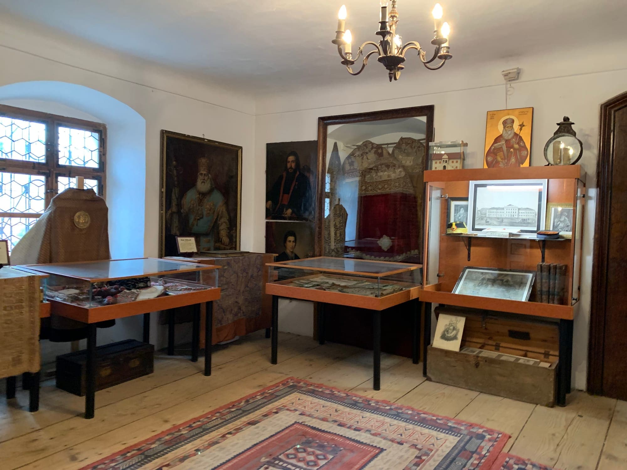 Sală a muzeului cu vitrine de lemn, portrete de ierarhi pe pereți, veșminte bisericești și un covor țesut