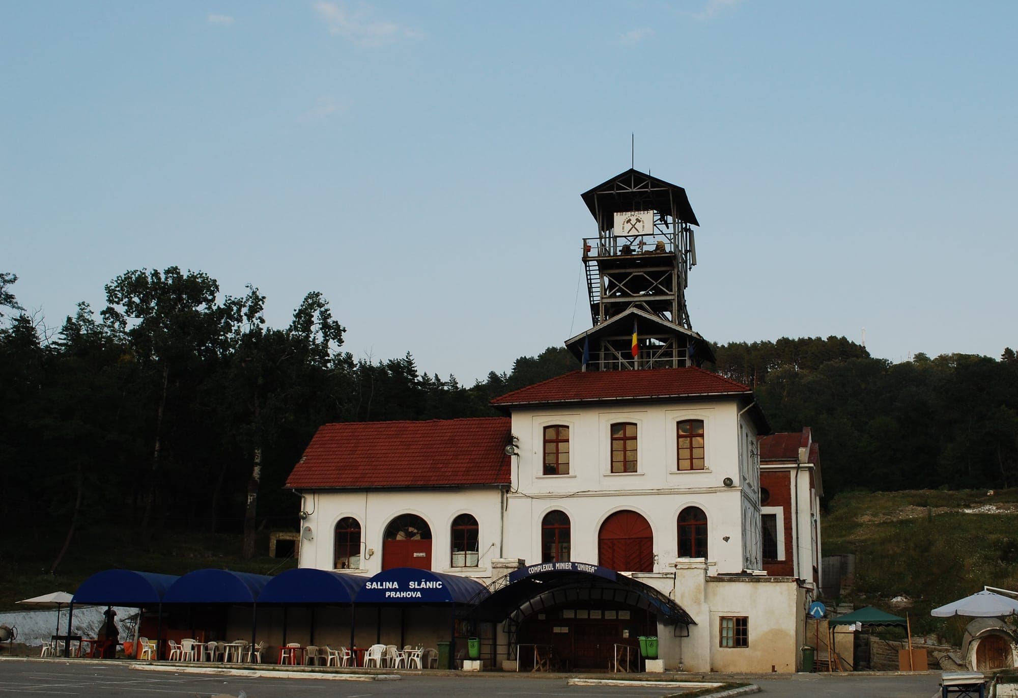 Clădirea albă a Complexului Minier Unirea, cu acoperiș roșu și turnul de lemn al puțului, la marginea pădurii