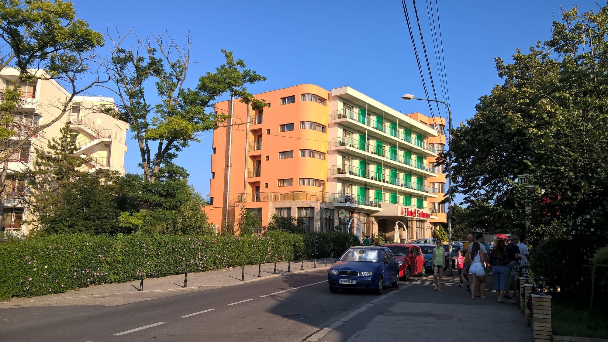Eine Straße im Badeort mit einem orangefarbenen Hotel mit grünen Balkonen, blühenden Hecken und Passanten auf dem Gehweg