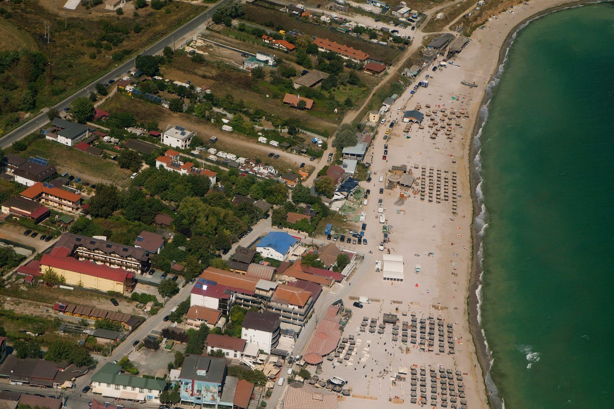 Vedere din aer spre satul Vama Veche și fâșia lată de nisip cu umbrele de stuf, cu marea verde-albăstruie în dreapta