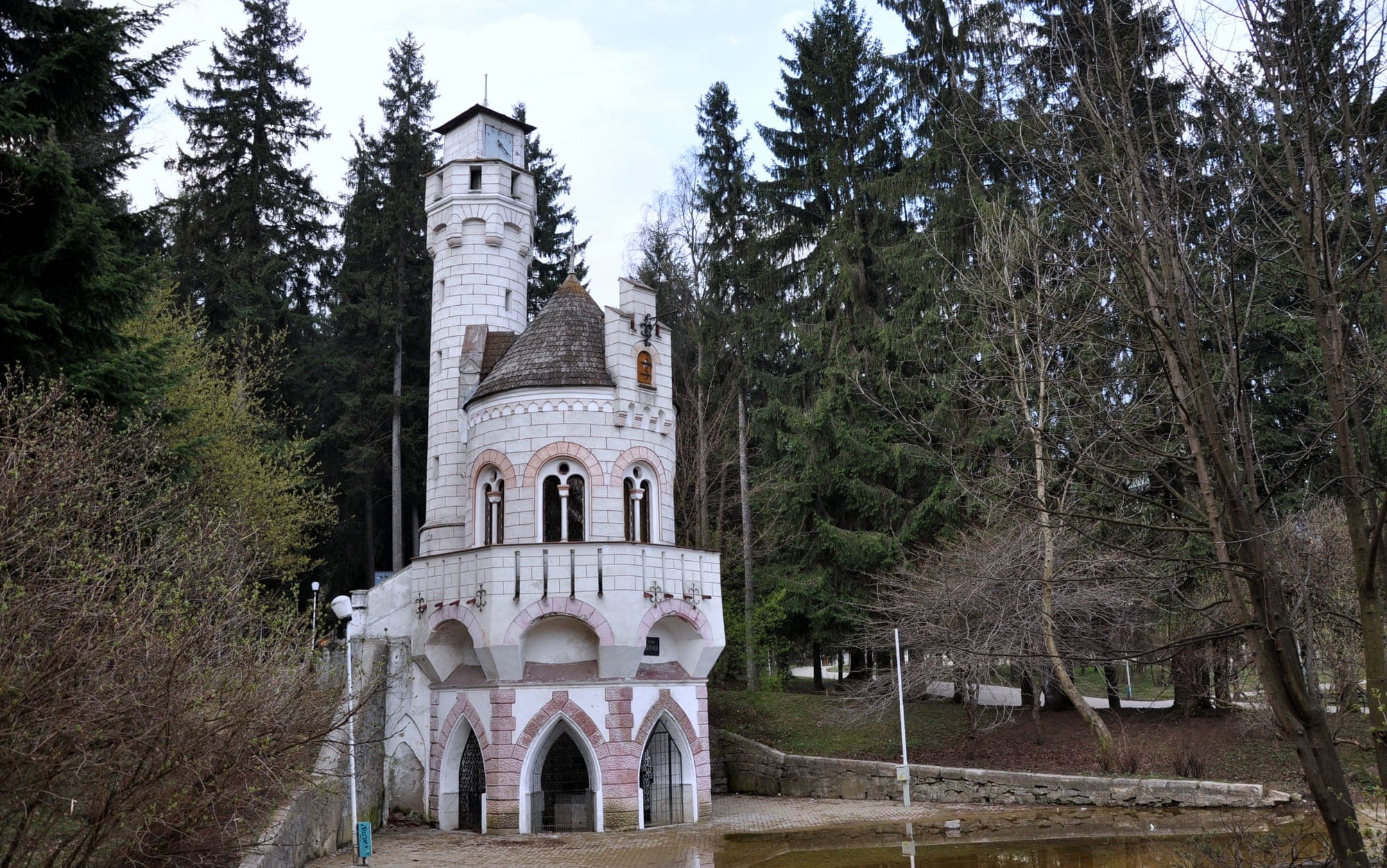 Pavilionul alb al Izvorului Sentinela, cu turn crenelat, arcade roz și acoperiș țuguiat, între brazii înalți din parcul băilor