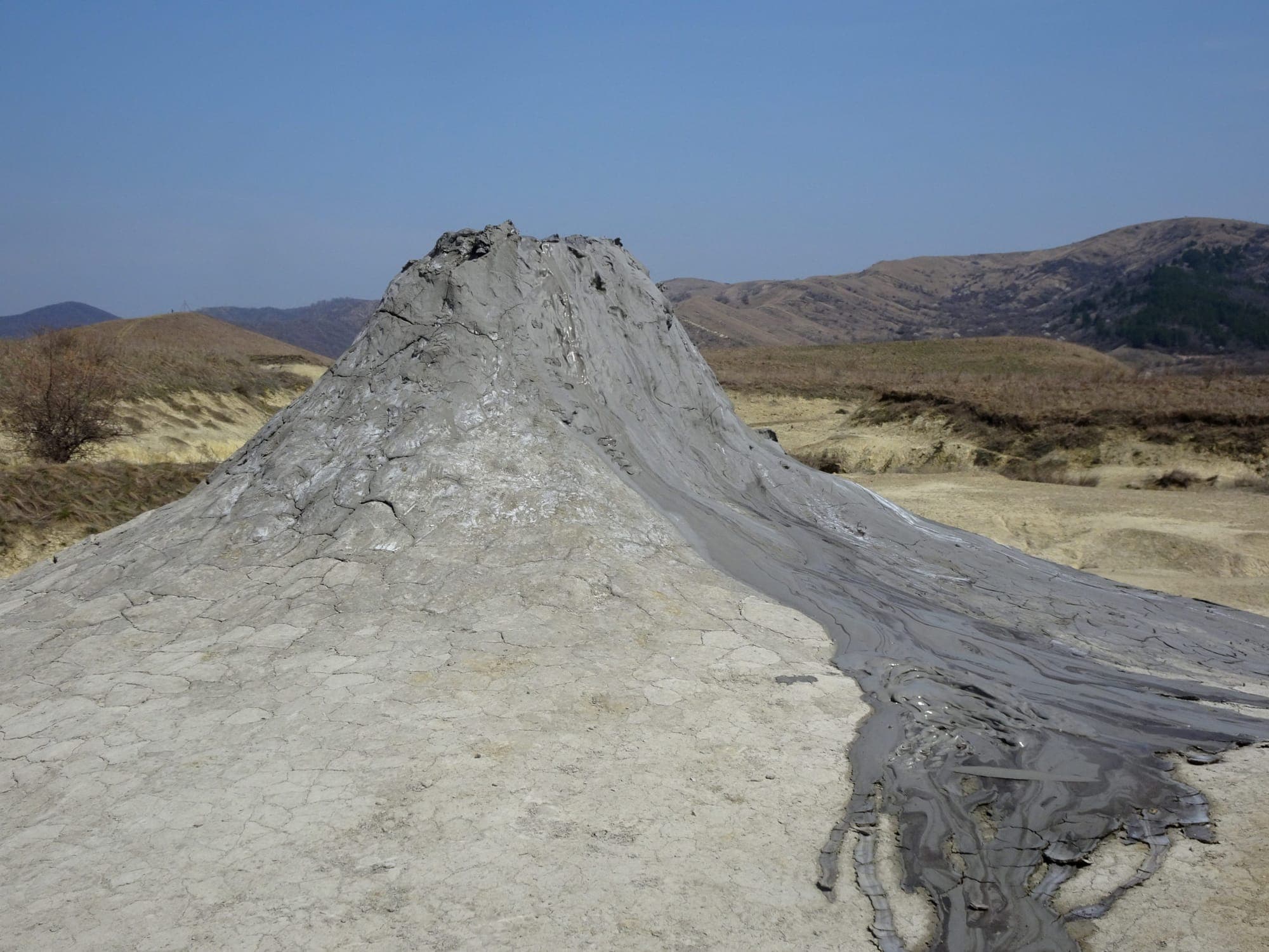 A tall cone of dried mud with the crater at its top and a stream of wet mud running down the slope, under a clear sky