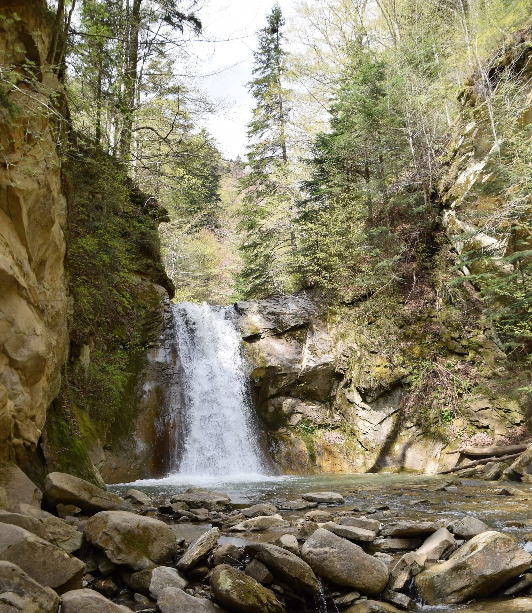 Cascada Cașoca căzând între pereți de gresie gălbuie și molizi, într-un bazin cu bolovani mari în prim-plan