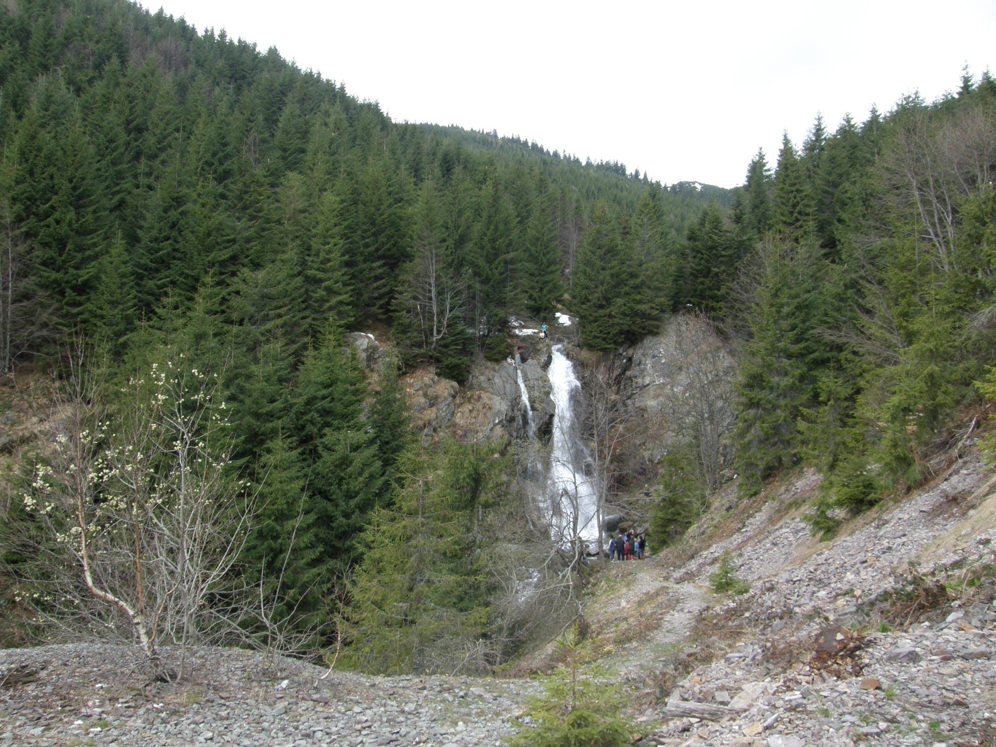 Cascada Vârciorog în fundul văii, între versanți cu molizi, cu grohotișul haldei de steril în prim-plan, primăvara