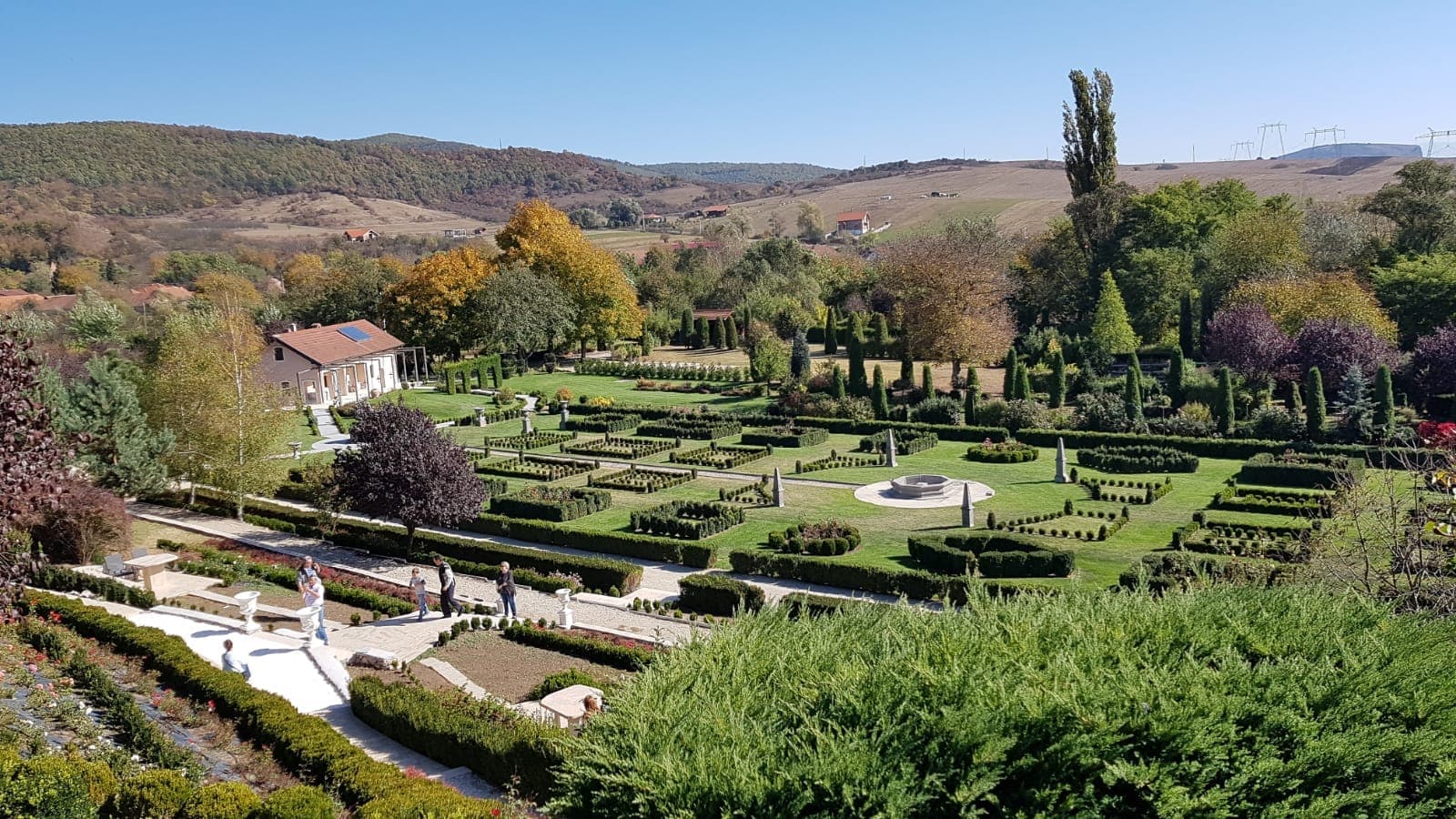 Vedere de sus a grădinii: parter cu garduri vii tăiate geometric și o fântână în centru, alei albe, o casă cu acoperiș roșu și dealuri toamna în fundal