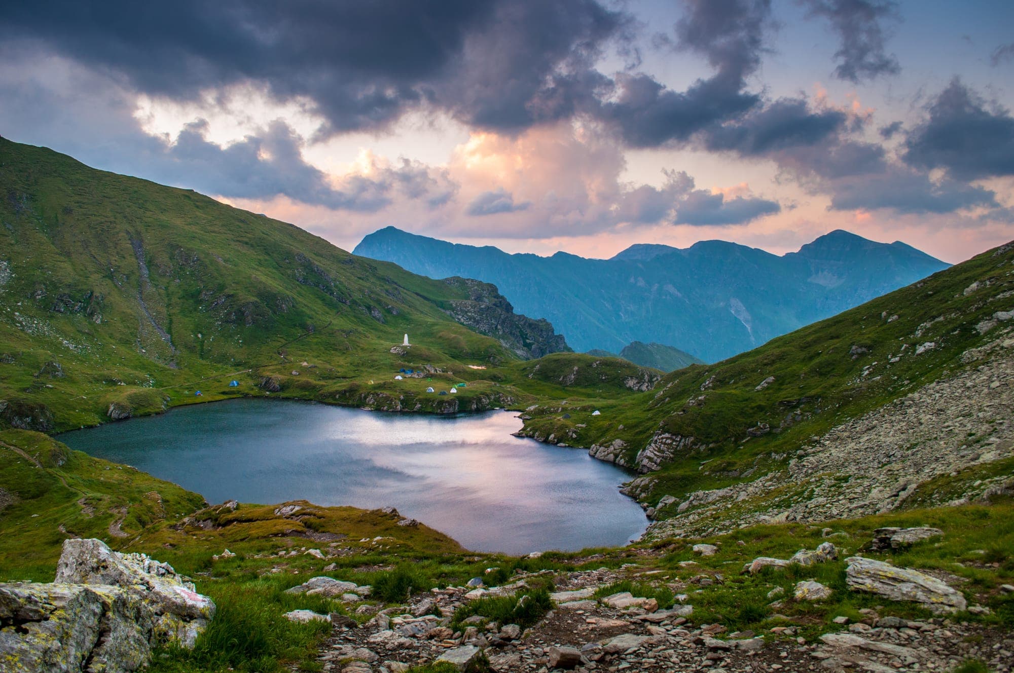 Lacul văzut de sus în lumina apusului, cu corturi și obeliscul alb pe malul opus și culmile Făgărașului în depărtare
