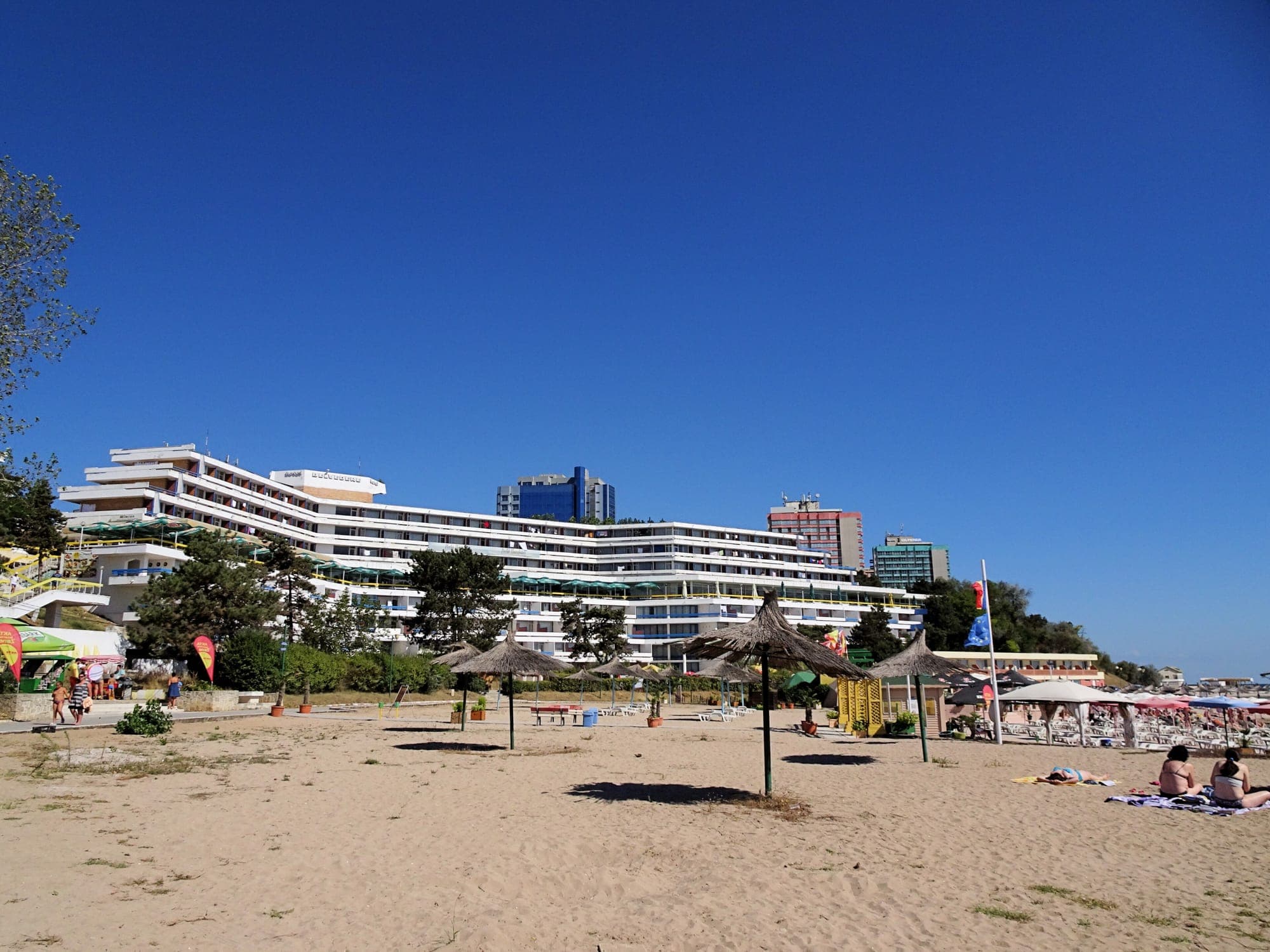 Hotel cu etajele retrase în trepte, la marginea plajei, văzut de pe nisipul lat cu umbrele de stuf, sub cer senin