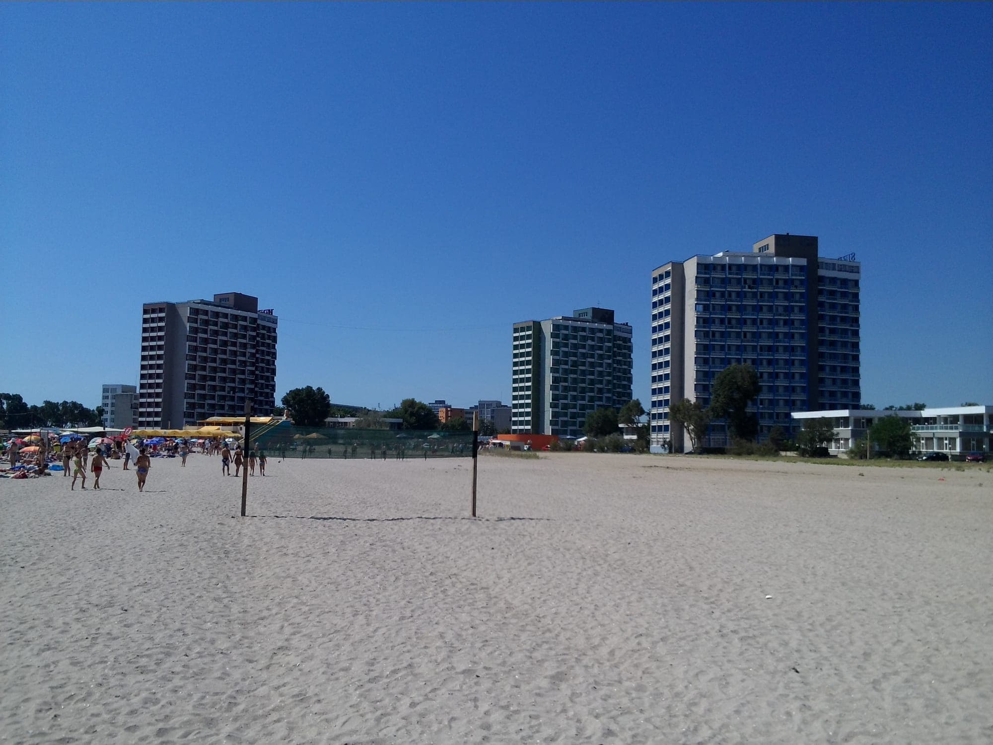 Drei hohe Hoteltürme vom weißen Sandstrand aus gesehen, mit einem Volleyballfeld davor und bunten Sonnenschirmen links